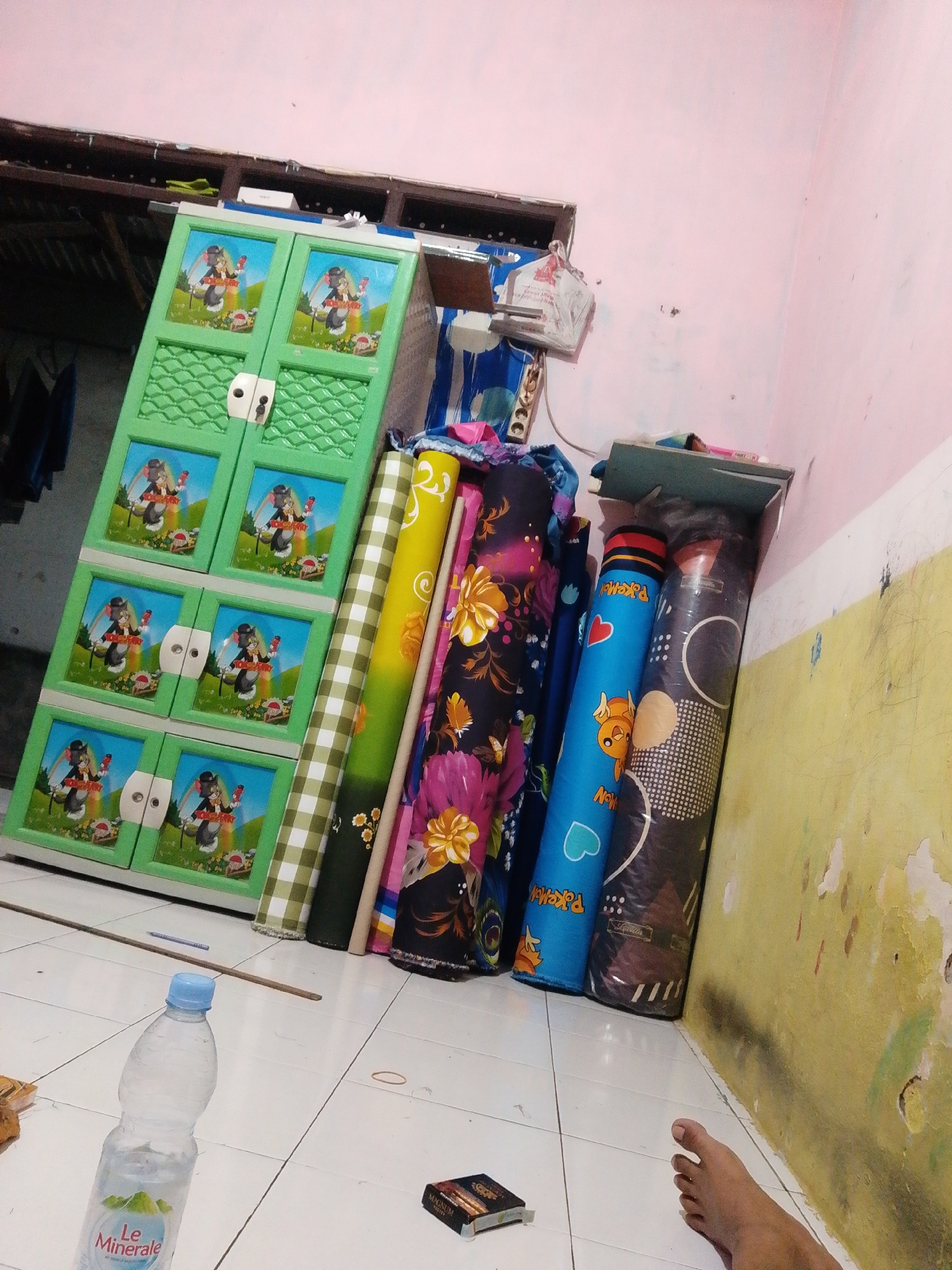 In the city of Bohar, Indonesia, a cluttered room contains a green plastic cabinet with cartoon characters on its doors, leaning against a light pink wall. Several rolled-up items, resembling mattresses or fabric rolls, are placed next to the cabinet and a small shelf. On the tiled floor, a plastic bottle of "Le Minerale" water and a pack of cigarettes are visible. A person's bare foot is also present in the lower right corner of the image. The environment appears to be an indoor residential space with signs of disarray.