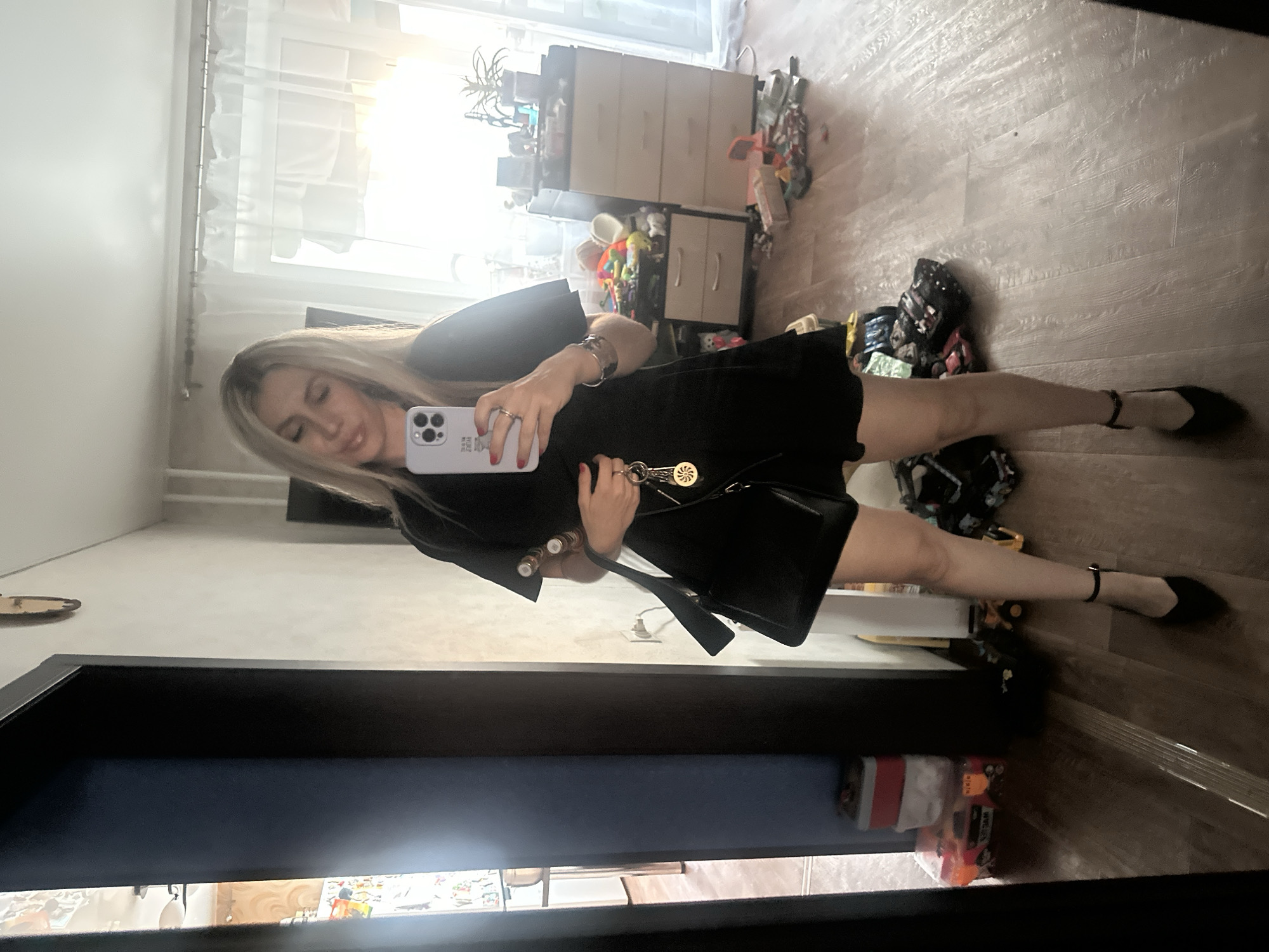 A woman is taking a mirror selfie in a room, possibly a bedroom or dressing area. She is standing and holding up her phone to capture her reflection. She has blonde hair and is wearing a black dress and black pointed-toe ballet flats. She is also carrying a black handbag. The room appears to be cluttered, with furniture, toys, and bags visible in the background. The lighting suggests it is daytime, with natural light coming from a window. The overall mood seems casual and informal. The location is identified as Novoaltaysk, Russia. No text is visible in the image.