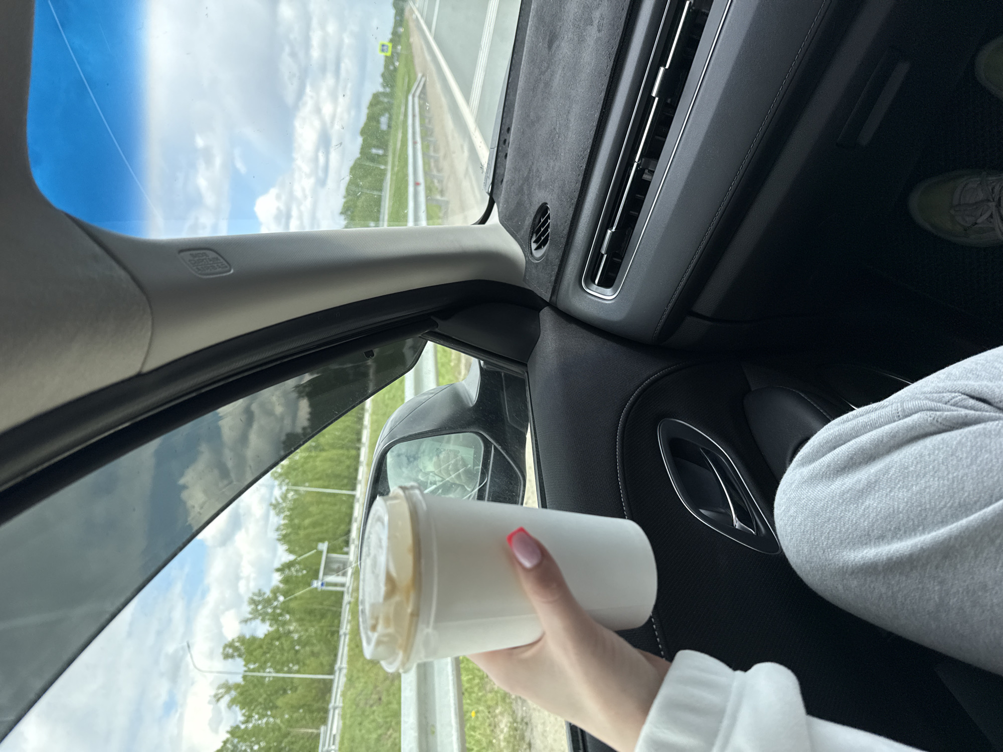 The image shows an interior view from inside a car, possibly on a highway or open road. A person is holding a white cup with a clear lid, presumably containing a beverage. Outside the car window, the scenery includes trees and what appears to be a highway with distant structures or signs. The sky is visible through the windshield, showing a partly cloudy day. The car's interior features a gray/black dashboard and trim. The person holding the cup has a hand with painted fingernails.