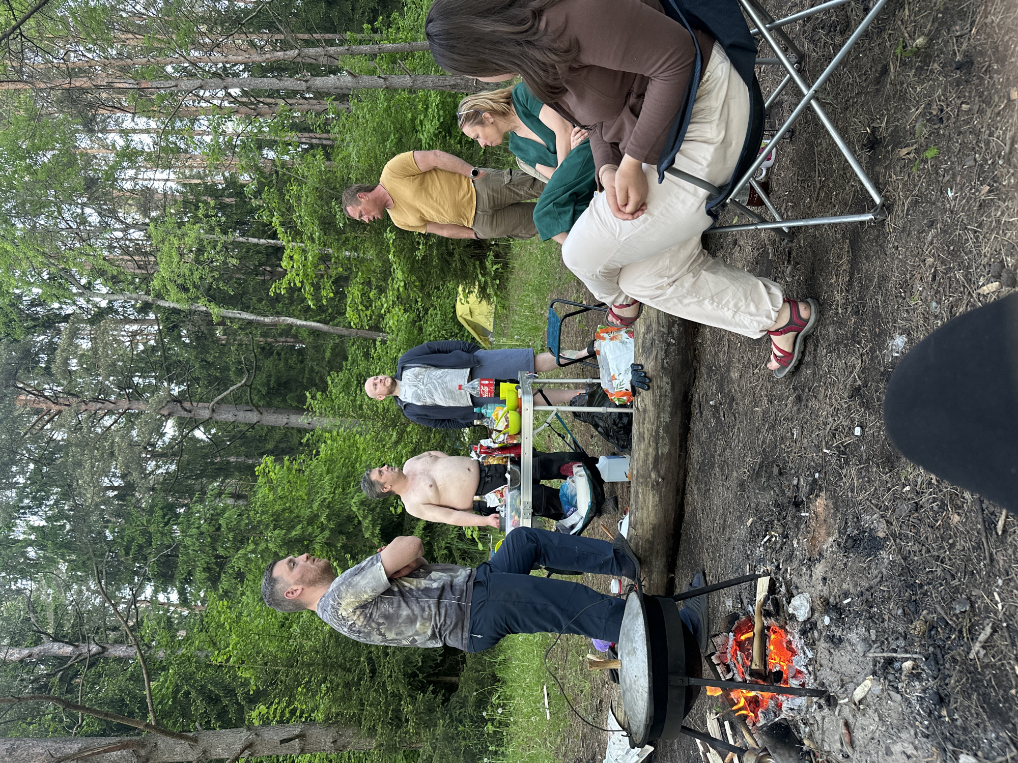 The scene shows a group of people gathered outdoors, likely in a wooded area, possibly near Юзуфоўскі сельскі Савет, Belarus, given the context. A fire is burning in a pit, supporting a large metal cooking pot or cauldron. A man stands beside the fire, possibly tending to the cooking. Several other individuals are present; some are seated on camping chairs, while others are standing nearby. A small table is set up with various items, including drinks and food. The environment consists of trees, undergrowth, and ground covered with dirt and debris. The image depicts a camping or picnic scene with multiple participants and objects related to outdoor cooking and relaxation.