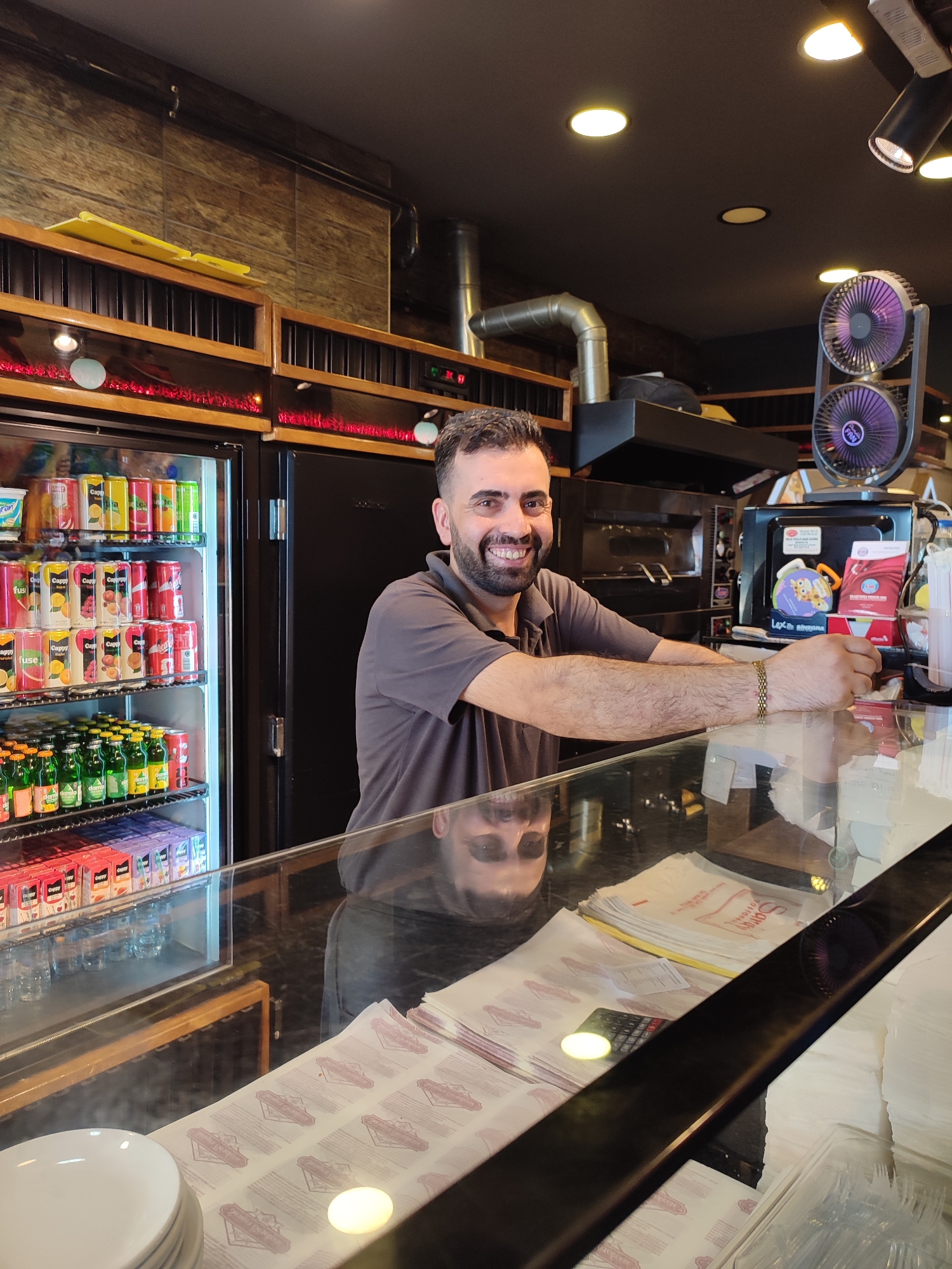 A man is standing behind a counter in what appears to be a food establishment, likely a pizzeria or a similar fast-food restaurant, located in Kadıköy, Turkey. The man is smiling at the camera. He has dark hair and a beard and is wearing a grey polo shirt. His arms are resting on the counter.
Behind him, a large refrigerator is filled with various beverages, including cans of soda and juice boxes. Above the refrigerator are shelves stocked with more items. Further back, a commercial oven and kitchen equipment are visible. The counter itself is made of glass and wood, and on it, there are stacks of paper, a calculator, and a small fan. The overall lighting suggests an indoor setting, possibly during the day or early evening.