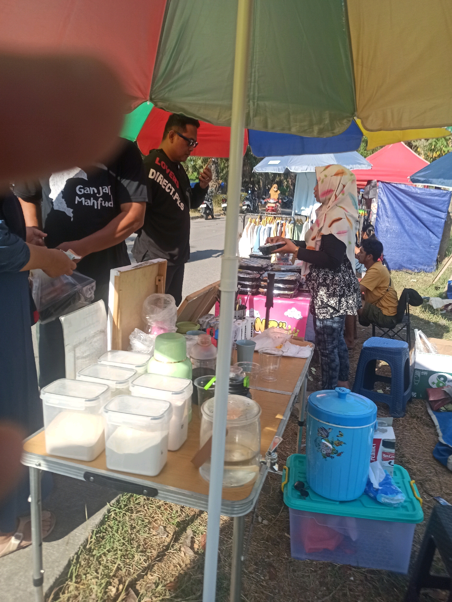 A medium shot captures an outdoor marketplace scene, likely in Telagamurni, Indonesia, under the dappled shade of several large, multi-colored umbrellas. In the foreground, a portable table displays several clear plastic containers filled with white powder, likely sugar or flour, alongside other small items such as green bowls and a clear glass dispenser with a spigot. A person in a blue long-sleeved shirt is partially visible on the left, interacting with someone in a black t-shirt that reads "Ganjar Mahfud." Another individual in a black t-shirt with "LOVERS DIRECT.PKS" printed on it stands in the middle, looking at their phone. Further back, a person wearing a white hijab is handing a black container to another person. Several racks of clothing are visible behind them, suggesting a retail area. A blue water cooler and various plastic storage bins are situated on the ground near the table. The ground is covered with dry grass and soil, indicating an outdoor setting. In the background, more tents and people are present, along with motorcycles. The lighting suggests it is daytime, with bright sunlight filtering through the umbrellas.