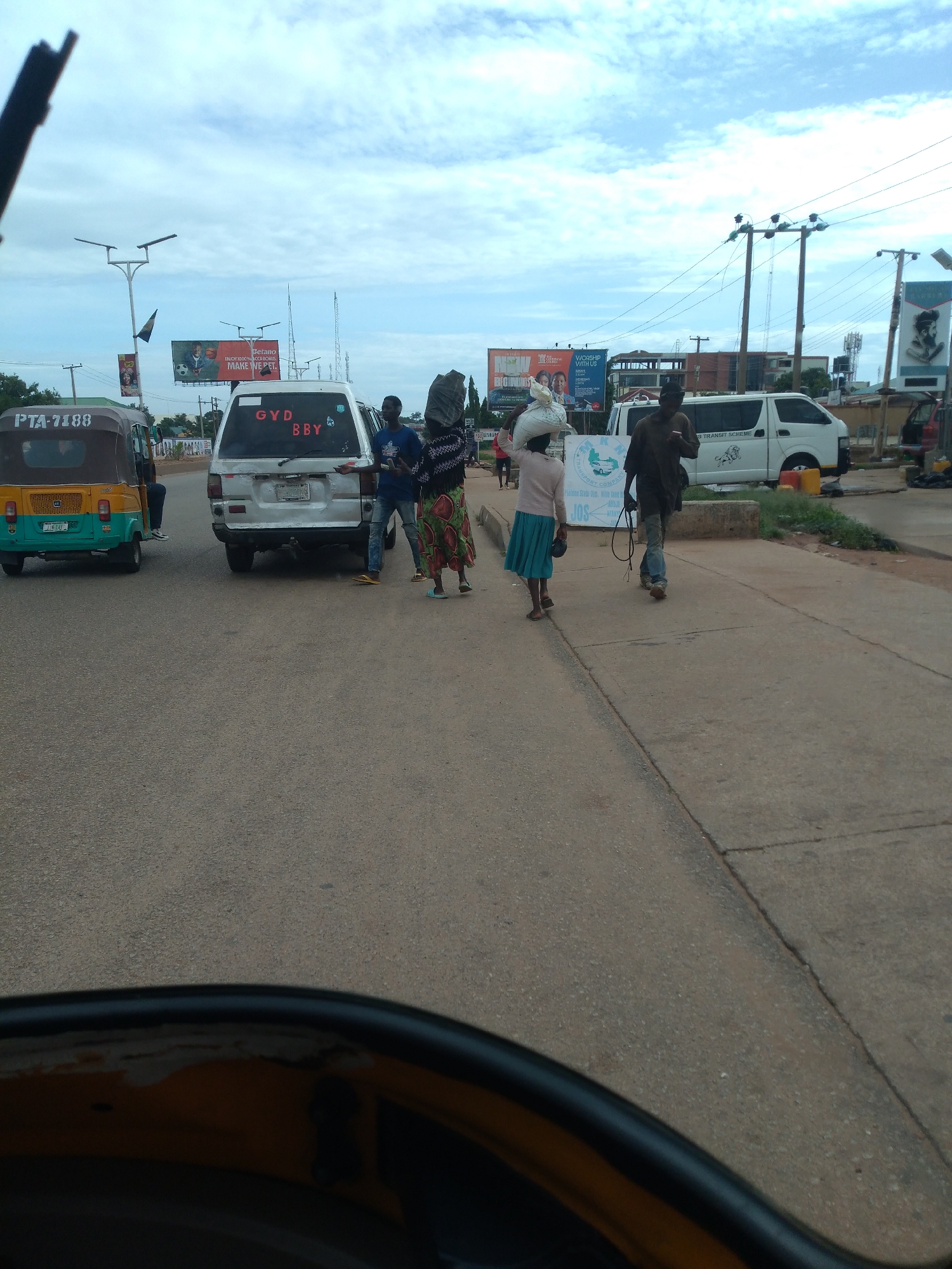 The image captures a detailed street scene in the city of Jos, Plateau State, Nigeria, during daylight hours under a cloudy sky. The perspective is from inside a vehicle, looking forward and slightly to the right onto an asphalt road with adjacent concrete sidewalks.
In the foreground, to the left, is a green and yellow three-wheeled auto-rickshaw (keke napep) with the license plate "PTA-7188" on its front, and "PLATEAU 43-43 JU" on a plate below it. Directly in front of the auto-rickshaw, a white passenger van is visible, featuring "GYD BBY" written in red on its rear window. A man in a blue t-shirt and jeans stands beside the driver's side of this white van, appearing to interact with someone inside or adjacent to the vehicle.
To the right of the first white van, a woman walks on the road, carrying a large, light-colored bundle on her head. She wears a long, patterned skirt with green, red, and yellow motifs and a dark top. Further to her right, a man walks on the concrete sidewalk, wearing a dark top, light blue jeans, and holding a thin, dark, flexible object in his right hand. Further left on the sidewalk, partially obscured, another person carries a white sack on their head and wears a light-colored top with a blue skirt.
The background features several elements:
* Multiple wooden utility poles with numerous overhead power lines crisscross the sky.
* Modern streetlights with dual lamp arms are mounted on tall poles.
* Various billboards and banners are present. One prominent billboard features football players and advertises "Betano - Enjoy the game. Play responsibly. Make W*n". Another banner further right displays a stylized portrait and clearly states "JOS". Another sign has "WORSHIP WITH US".
* In the mid-ground, a second white minivan with a lion logo and "TRANSIT SCHEME" text on its side is parked or moving slowly. Two yellow plastic jerricans are visible on the sidewalk near this van.
* Low-rise buildings and some vegetation line the street further into the background. Concrete barriers separate parts of the road from the sidewalk.
The scene depicts typical urban activity, with pedestrians, vehicles, and commercial advertisements common to Nigerian cities.