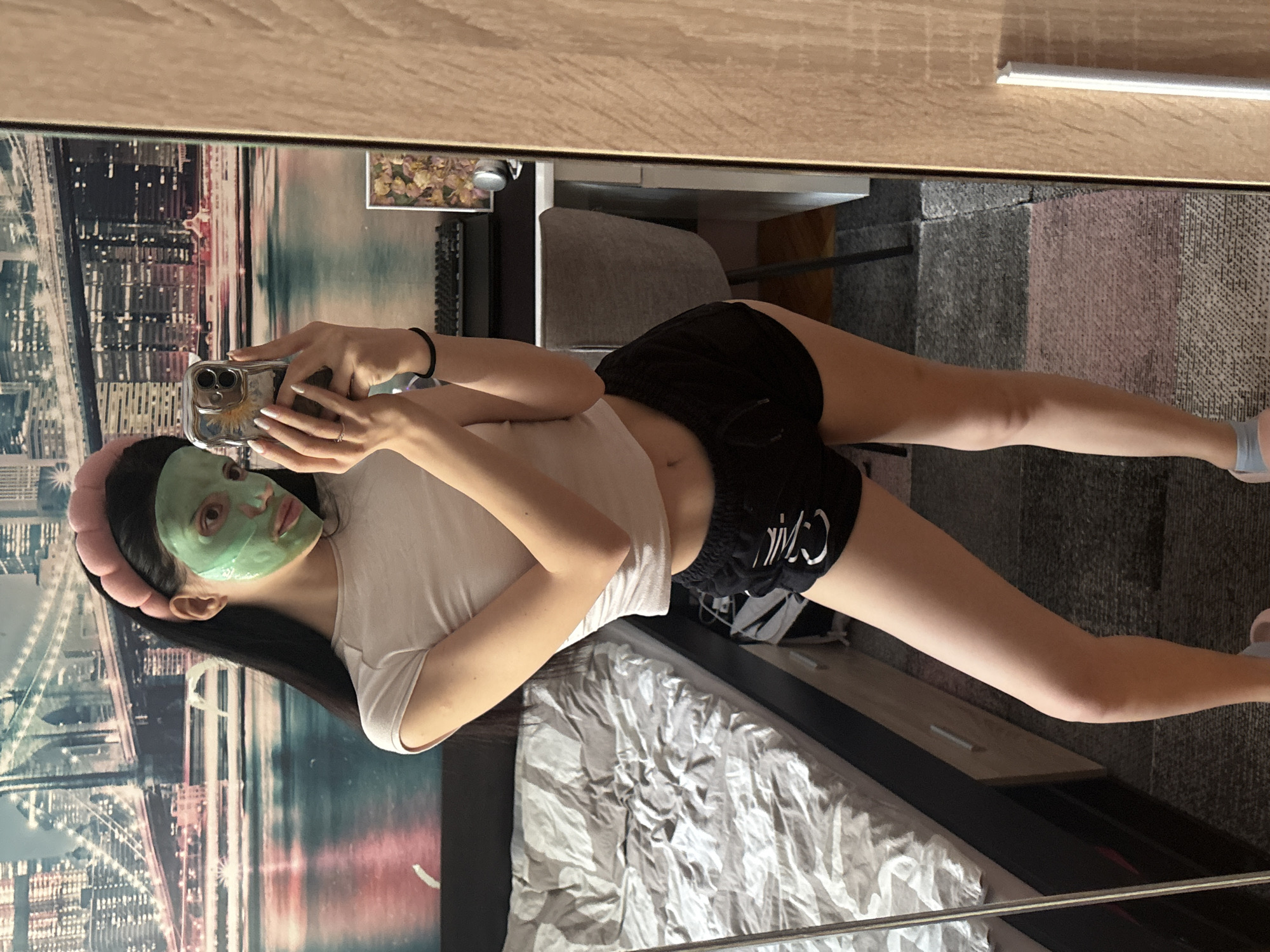A woman is lying on a bed, taking a selfie in a mirror. She has a green facial mask and a pink headband. She is wearing a white t-shirt and black shorts. The bed has a white comforter. The room contains furniture like a desk, a chair, and a drawer. In the mirror’s reflection, there is a city scene in the background, looking like an art print. The location in the city of Sofia, Bulgaria, cannot be determined from the provided image.