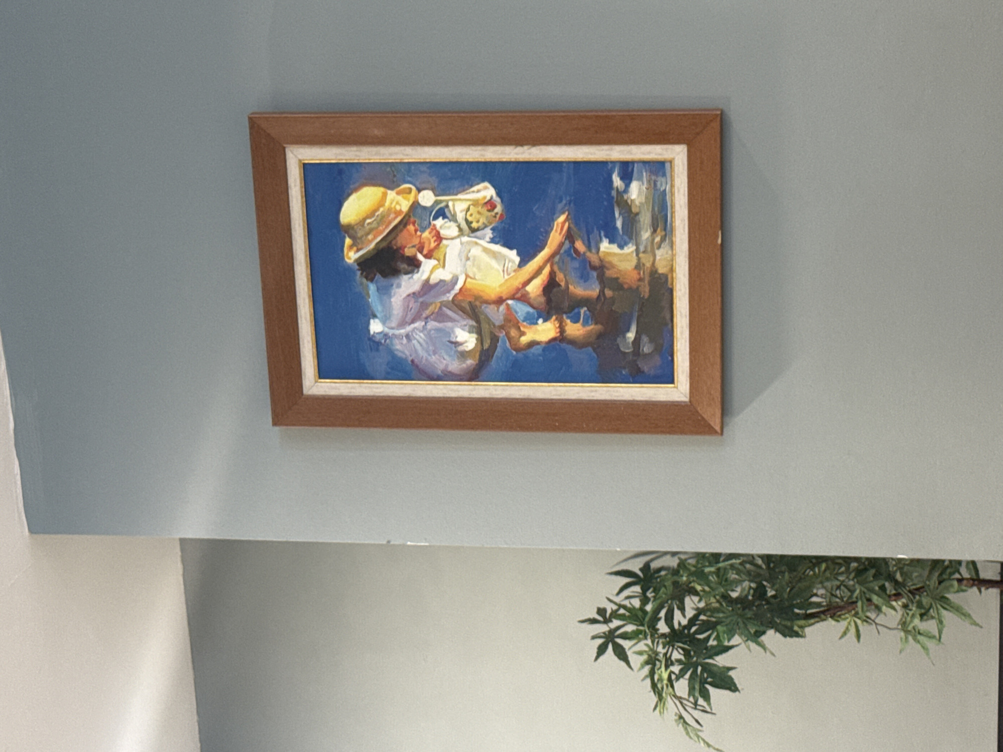 A framed oil painting depicts a young person, possibly a child, wearing a yellow sun hat and a light-colored dress, crouching down and reaching towards a body of water. The figure is reflected in the water, which appears to be a vibrant blue. The painting is hung on a light blue wall. To the left of the painting, partially in the frame, is a potted plant with green, leafy foliage. The overall scene suggests an indoor setting, possibly a home or gallery in the city of Aksu, Turkey.