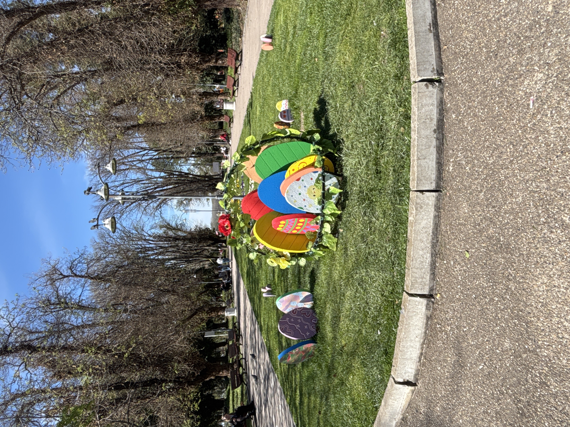 The image depicts a park in Silistra, Bulgaria. In the foreground, there is a large arrangement of colorful, decorative Easter eggs placed on a patch of green grass. Some smaller, scattered egg decorations are visible further away on the grass. To the right, there is a concrete pathway. On the left, there is a tree-lined avenue with benches and streetlights. Bare trees without leaves are visible, suggesting it may be early spring. The sky is blue with minimal clouds. In the background, there are other park elements, including more trees and possibly buildings. There are indications of people in the background.