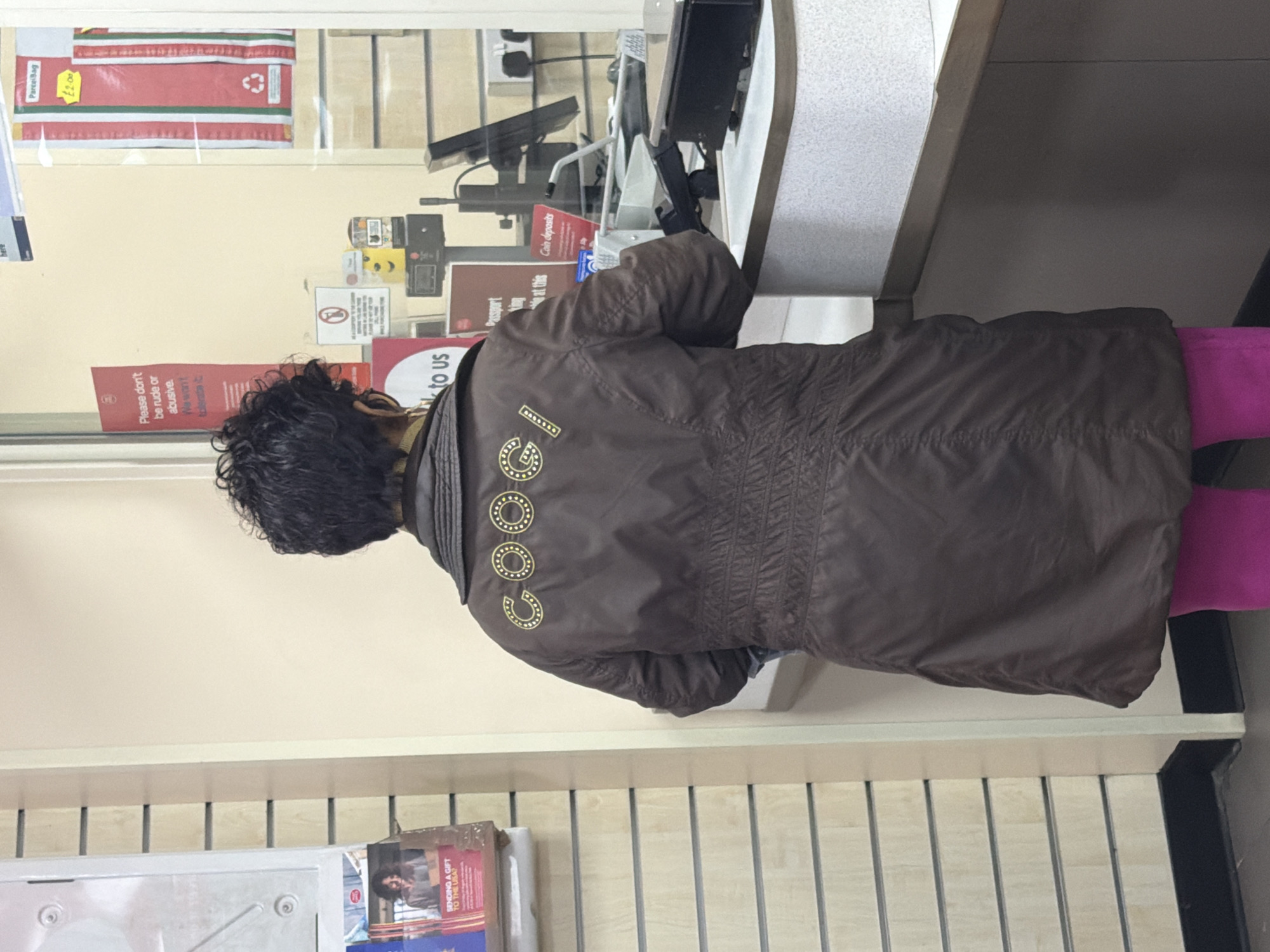 A woman with curly black hair, wearing a brown jacket with the inscription "COOC," is standing at what appears to be a post office counter. She is also wearing magenta-colored tights. The jacket has gold-colored circles on the back. The post office counter has a screen and other technological equipment on it. A glass partition separates the customer area from the employee area. Various signs are visible behind the glass, including notices stating "Please don't be rude or abusive," "Passport," "Coin deposits," and a promotional ad that states, "SENDING A GIFT TO THE USA?" A "Parcel Bag" with a price tag of "£2.00" is displayed on the glass. A slatted wood-like paneling can be seen on the wall. The location is likely within the city of London, United Kingdom, based on the presence of signage that mentions the UK post system.
