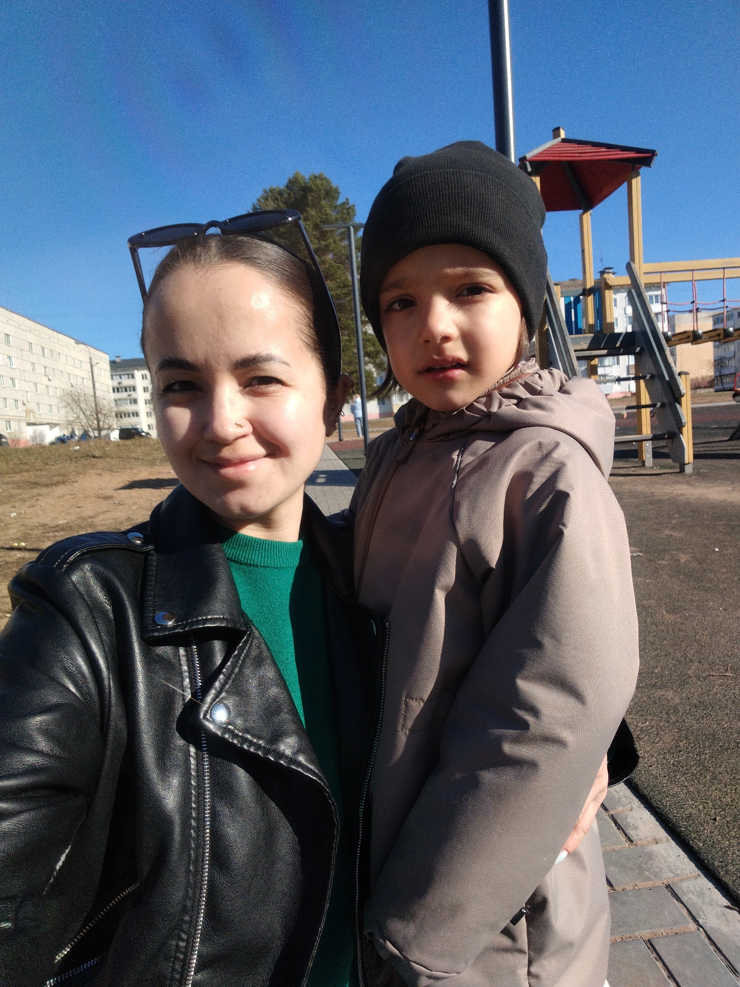 A bright, sunlit outdoor portrait captures an adult woman holding a young child at a playground in Noviy, Russia. The adult, positioned in the foreground on the left, appears to be taking a selfie. She has dark hair pulled back, sunglasses resting on her head, a subtle nose piercing, and a gentle smile directed at the camera. She is dressed in a black leather jacket over a green, ribbed sweater.
Cradled in her arms is a child, likely between four and seven years old, wearing a black beanie and a light brown or taupe hooded jacket. The child has a neutral expression while looking towards the camera.
The background reveals a clear blue sky and strong sunlight casting distinct shadows. Elements of an urban environment are visible, including a colorful playground structure with yellow, red, and blue components, and several multi-story apartment buildings in the distance. The ground features bare earth and a paved walkway, with a tree displaying some foliage. No visible text is present in the image. The scene suggests a sunny day, possibly in early spring or late autumn given the ground cover.