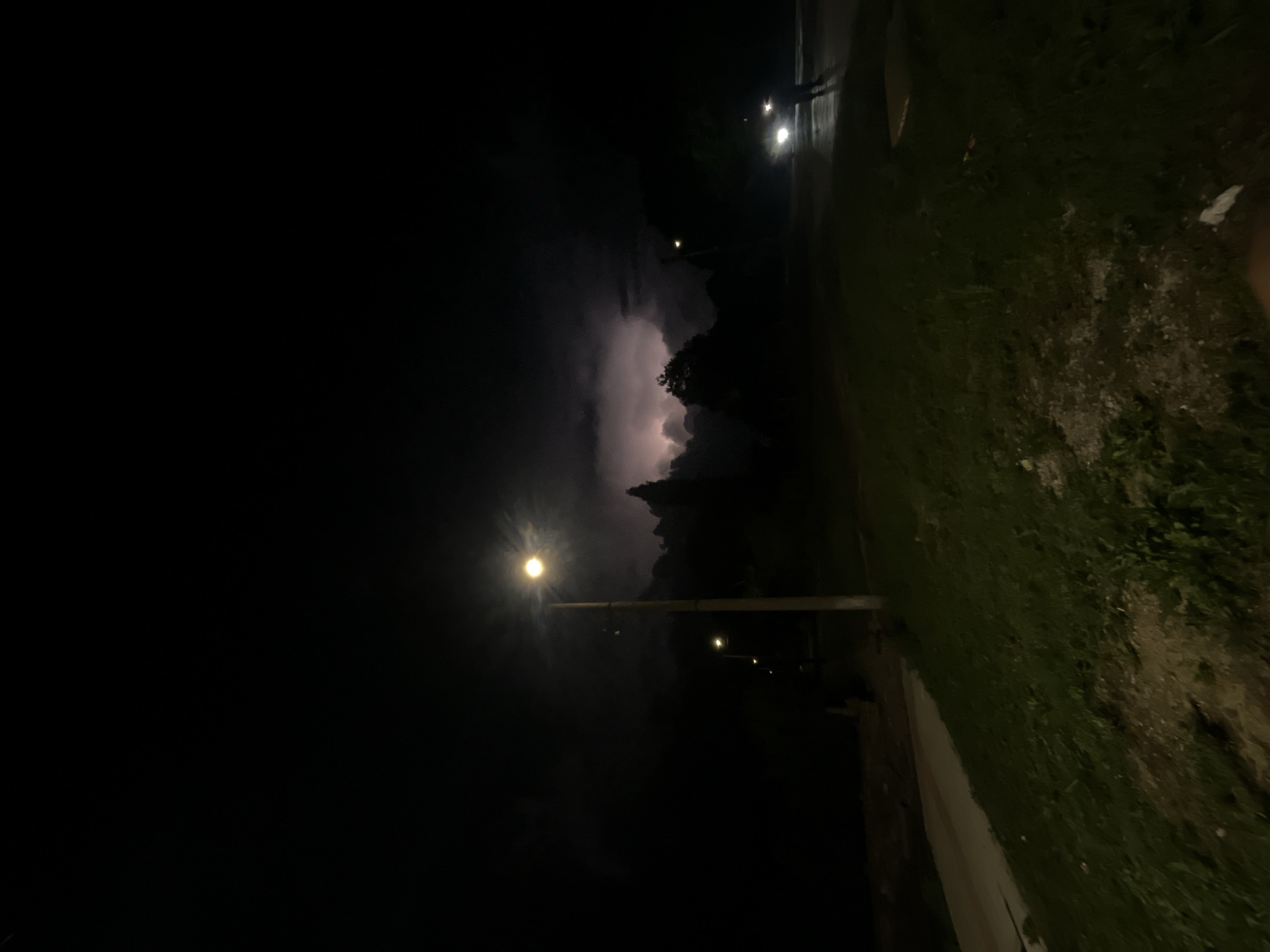 Night shot in Lozarevo, Bulgaria. A streetlight illuminates the lower left portion of the image, revealing a sidewalk and grassy area to the right. Further in the distance, a cluster of trees and clouds suggests a storm with lightning present. On the street, to the right of the trees, headlights of a car are visible.