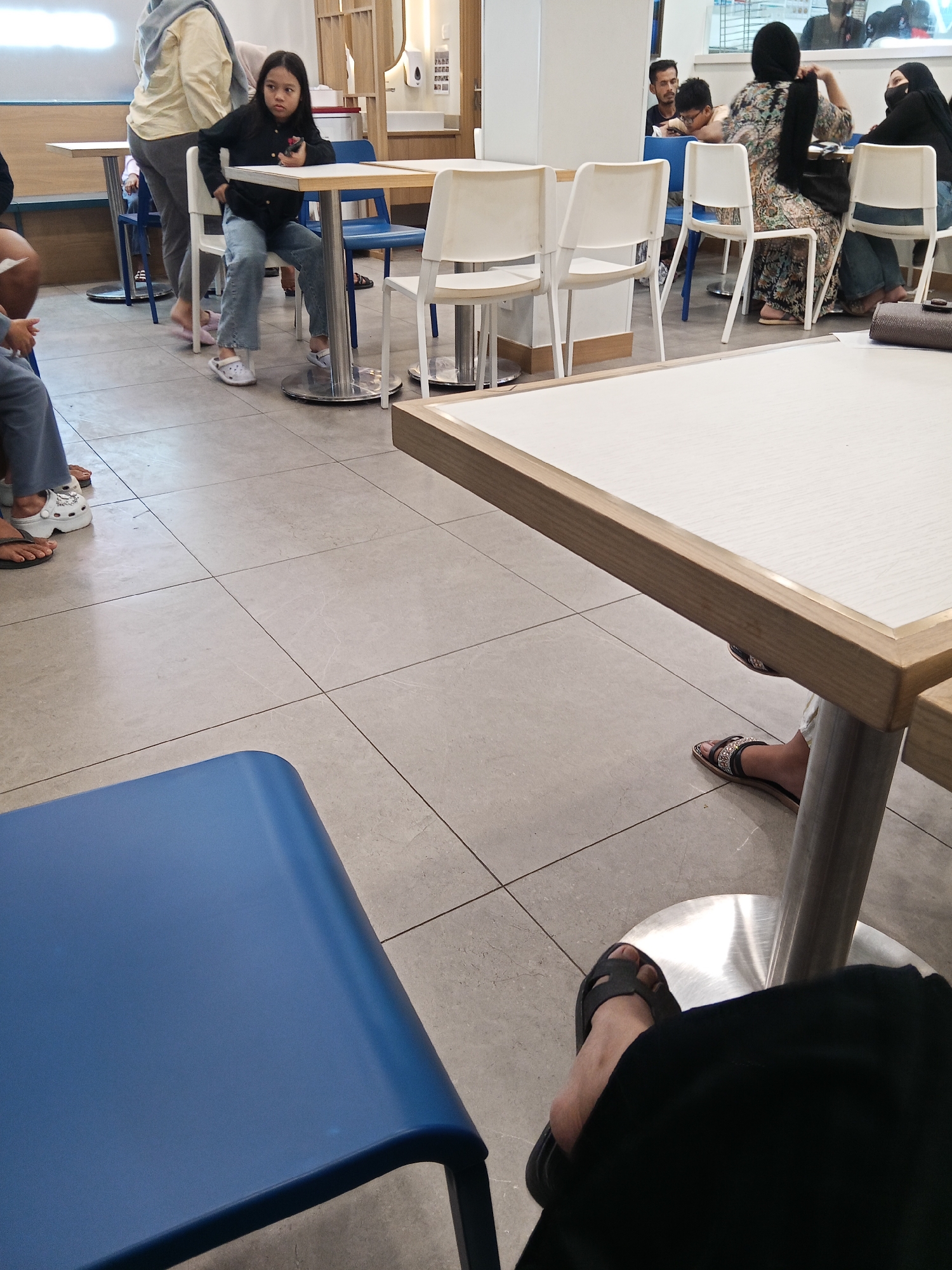 The image depicts an indoor scene, likely a cafe or restaurant, with a focus on a young girl sitting at a table. She is wearing a black long-sleeved shirt and blue jeans. In the background, several other individuals are present, some seated at tables and others standing. The environment features tiled flooring, metal-legged tables, and chairs. The lighting appears to be artificial, creating a bright atmosphere. There are no discernible landmarks or signs that would pinpoint the specific location in Lubuklinggau, Indonesia.