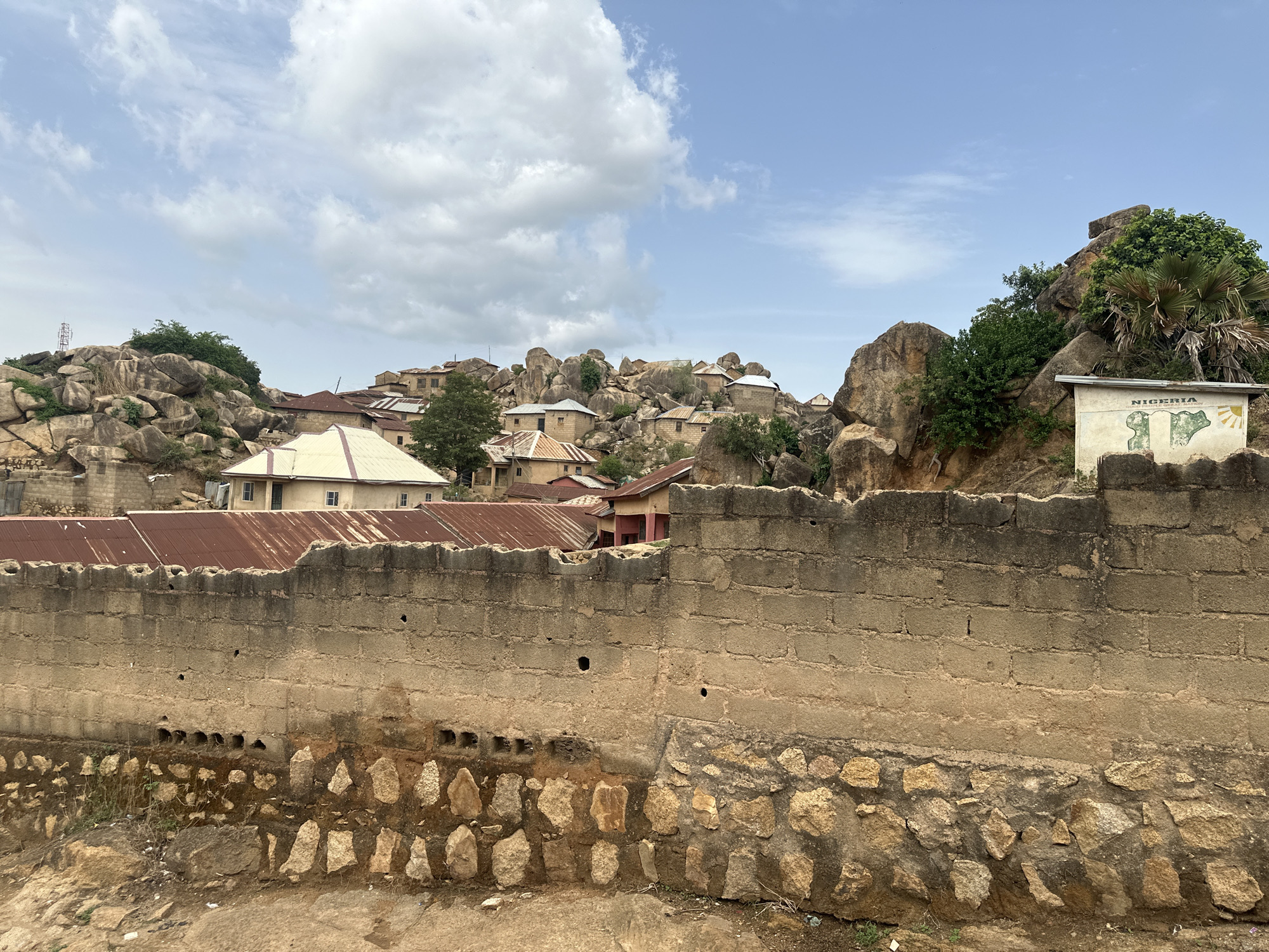 The scene depicts a village in Jos, Nigeria. In the foreground is a weathered wall comprised of stone and brick. Behind the wall are structures with corrugated metal roofs, interspersed with large boulders and vegetation. A sign with a map of Nigeria is affixed to a structure near a rocky outcrop on the right. The sky is partly cloudy.
