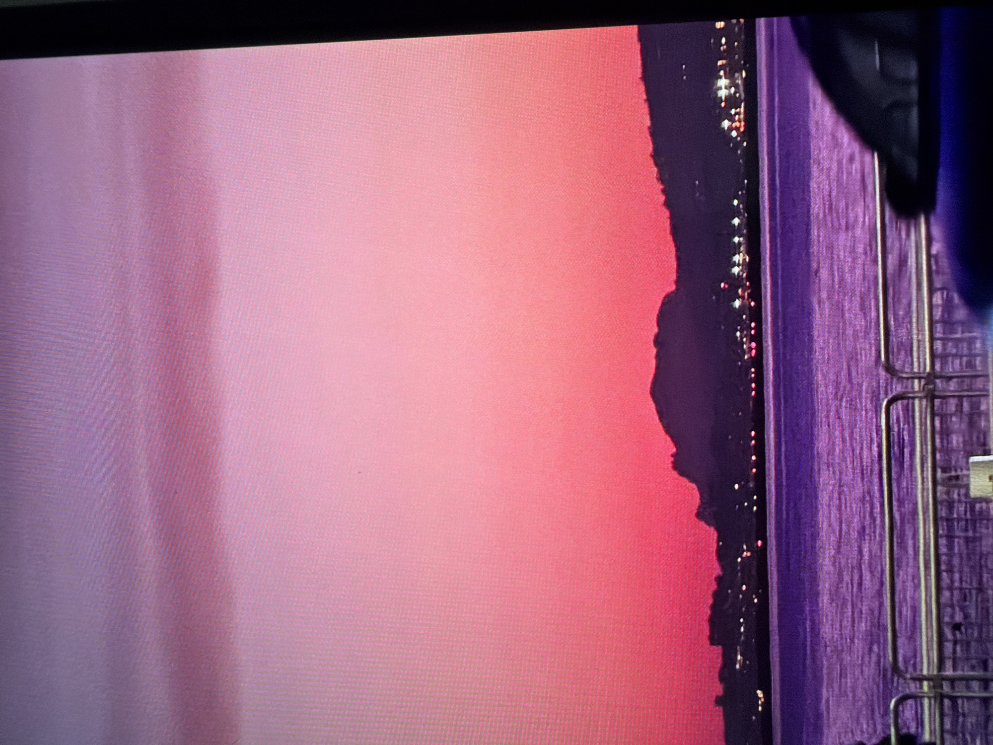 This is a photograph taken in Sant Antoni De Portmany, Spain, featuring a vibrant sunset over the water. The sky is awash in a gradient of pink and purple hues, transitioning from a lighter lavender on the left to a deeper, more intense pink towards the right. On the far right of the image, the purple water reflects the colors of the sky.
Silhouetted against the colorful sky is the dark outline of a town or coastline. Small, bright lights dot the edge of this silhouette, indicating that it is nighttime or dusk and the town is illuminated. The contrast between the dark silhouette and the brightly lit lights against the sunset creates a dramatic effect.
To the right of the silhouette, a textured surface, possibly a building or wall, is visible in shades of purple. In the foreground, a railing with vertical metal bars is present, suggesting a viewpoint from a balcony, promenade, or structure overlooking the scene. The overall mood is serene and visually striking, capturing the beauty of a Mediterranean sunset. The time of day is clearly evening, and the weather appears clear, allowing for a unobstructed view of the sunset. There is no visible text in the image.