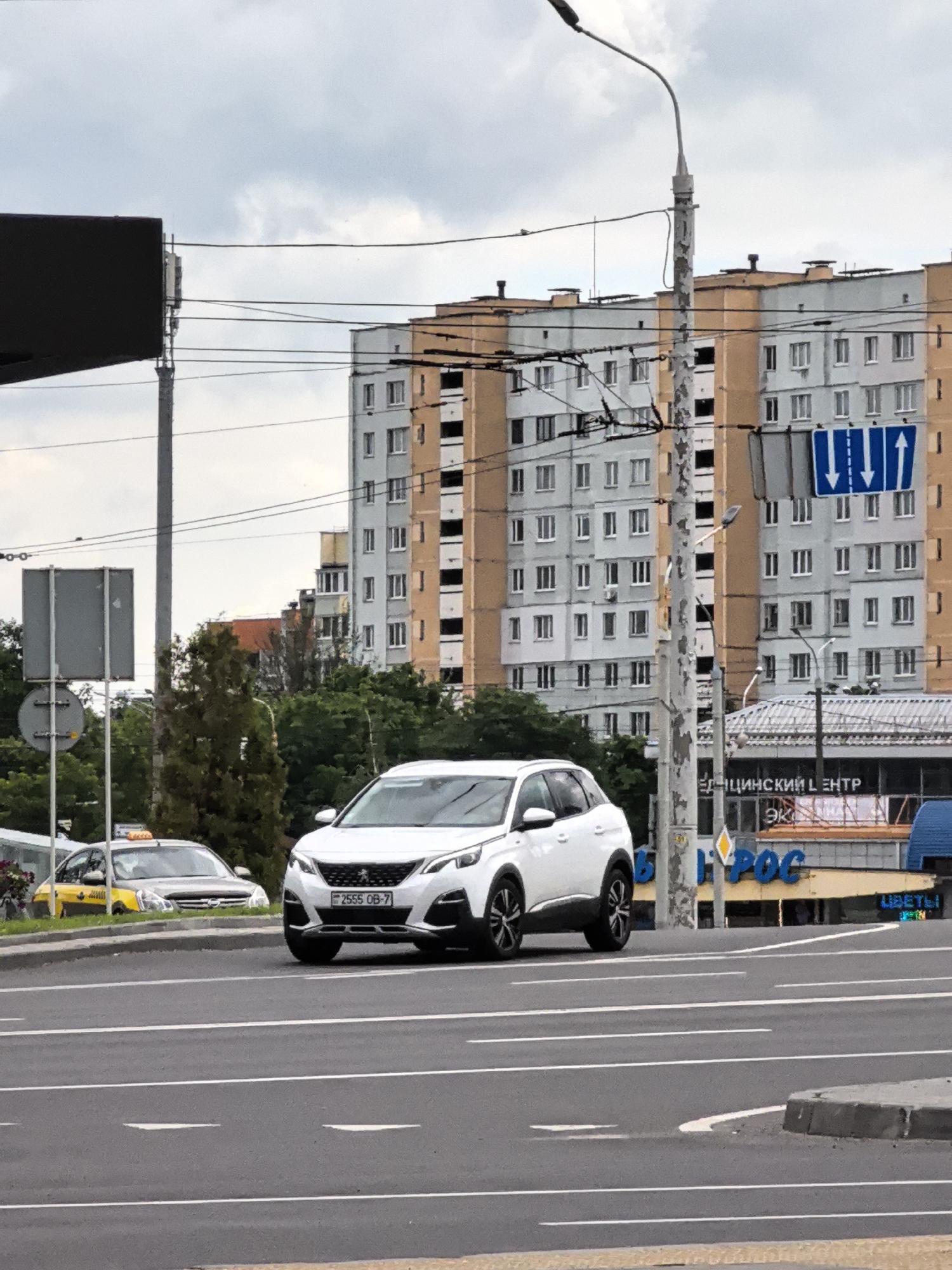 A white Peugeot 3008 SUV is driving on a paved road in an urban environment. The car, with license plate "2555 OB-7", is in the foreground. In the background, there are multi-story apartment buildings, trees, a traffic light, and a taxi cab. The sky is overcast.