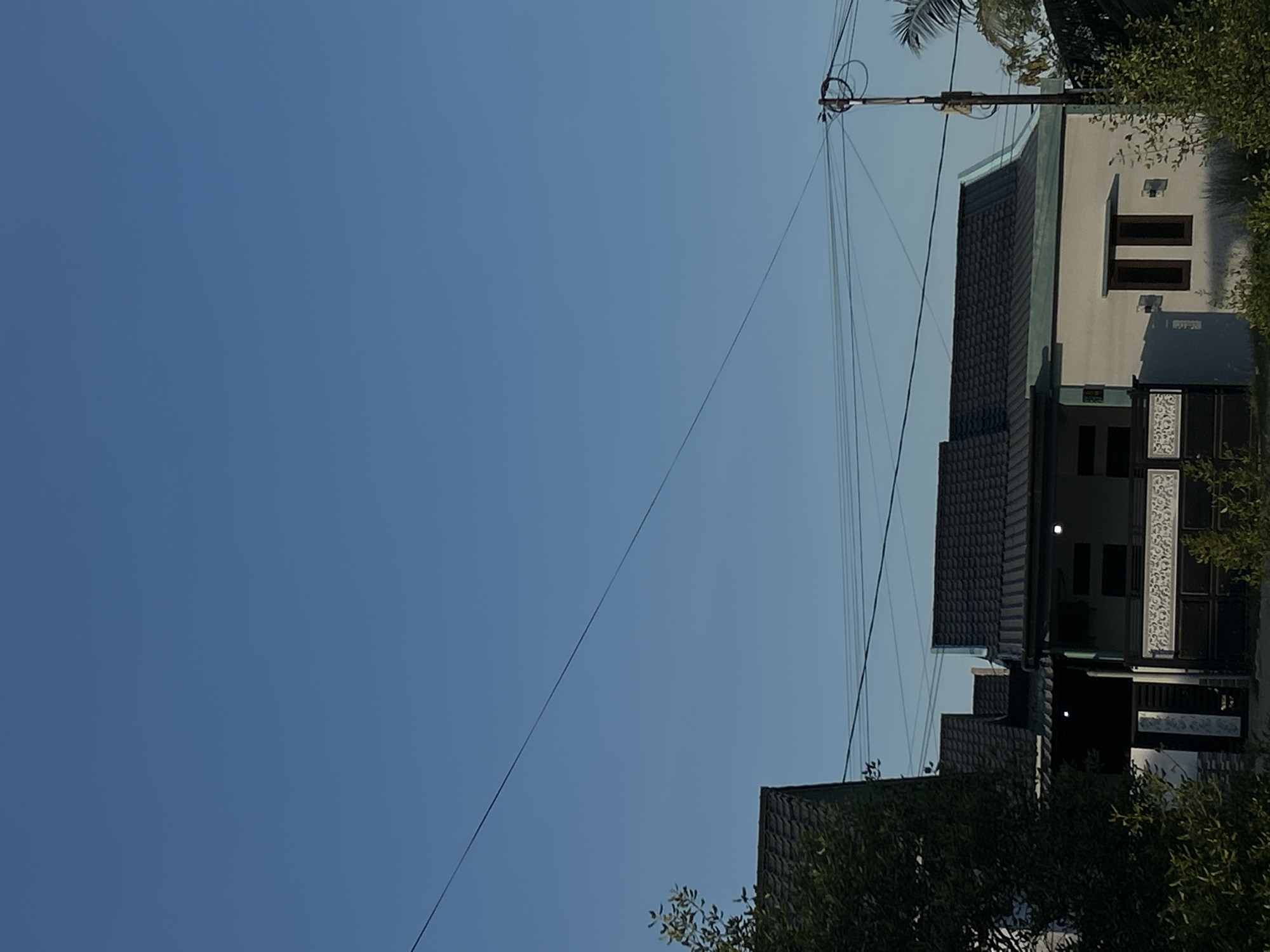 The image captures a view from Lhokseumawe, Indonesia, focusing on a clear blue sky occupying the majority of the frame. Power lines stretch across the sky, connecting to a utility pole. Adjacent to the pole, a multi-story building with a dark, gabled roof is visible. The building's facade is painted a light color, with dark-framed windows and decorative panels. The right side of the frame is partially obscured by trees and foliage.
