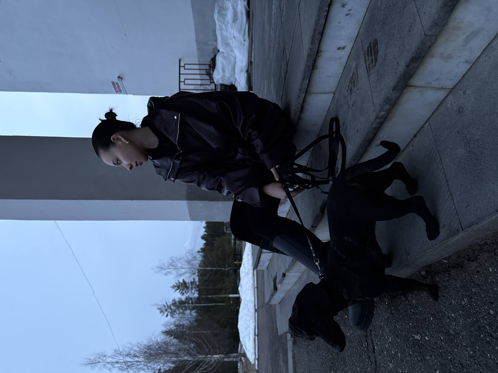 A woman sits on a stone step next to a black dog on a leash. The woman has dark hair pulled back in a bun and wears a dark jacket and leggings. The dog stands next to her, facing to the right.
The steps are part of a stone building, and behind the building are trees and a cloudy sky. There is snow on the ground behind the steps.
The location is in Noviy, Russia.