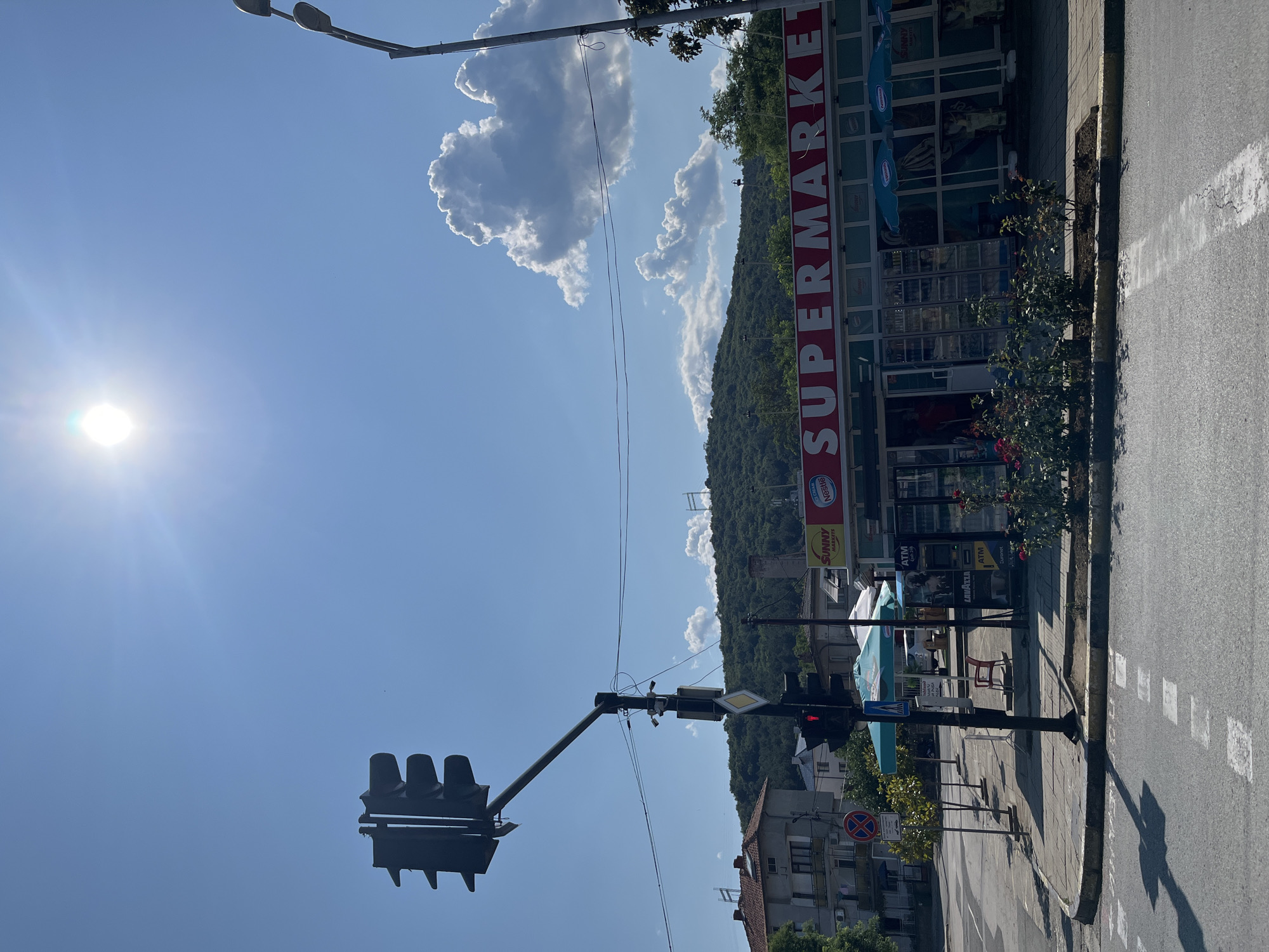 The image shows a street scene in Obzor, Bulgaria, on a clear, sunny day. The sun is bright, indicating it's likely daytime. In the foreground, a traffic light is visible with its red light illuminated, suggesting traffic is stopped. Adjacent to the traffic light is a street sign with a diamond shape. The scene then moves to the right, revealing a supermarket with a large "SUPERMARKET" sign in red lettering. The supermarket has large glass windows and doors, with several displays visible inside. Some umbrellas and seating are set up outside the supermarket, suggesting a small outdoor area. Beyond the supermarket, a densely forested hill rises, with some clouds in the bright blue sky. A street pole with lights is also visible. The overall impression is a typical street view of a town on a bright, pleasant day. There is also text on signs for "ATM", "Nestle", and "Sunny Markets".