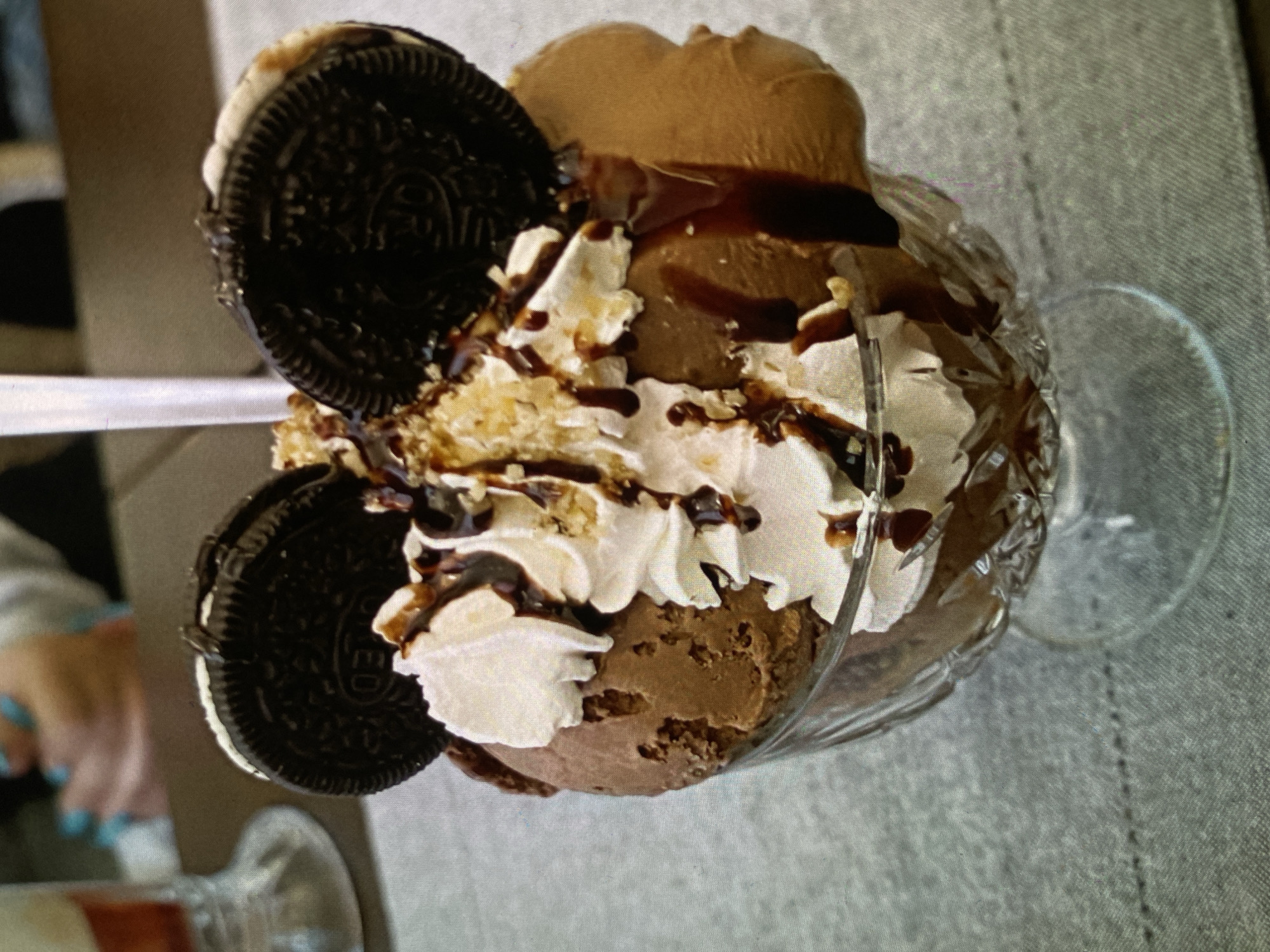 An overhead, slightly angled shot captures a decadent dessert presented in a clear, ornate, stemmed glass, resembling a sundae. The dessert features several scoops of dark brown ice cream, likely chocolate or coffee flavored. It is lavishly topped with generous swirls of white whipped cream, over which a rich, dark chocolate syrup is artistically drizzled. Light-colored, crushed toppings, possibly cookie crumbs or nuts, are sprinkled across the whipped cream and ice cream. Two whole Oreo cookies, with their distinctive dark chocolate wafers and "OREO" branding clearly visible, are decoratively placed on opposing sides of the dessert. A white utensil, appearing to be a spoon or thick straw, is partially inserted into the dessert from the left side.
The glass rests on a light gray, textured surface, possibly a tablecloth or tabletop. In the softly blurred background to the lower left, a person's hands with light blue fingernail polish are faintly visible, suggesting human presence nearby. The overall scene depicts an appetizing and indulgent treat. The time of day and specific indoor location are not visually identifiable from the image itself, though the provided context places the location in Bankya, Bulgaria.