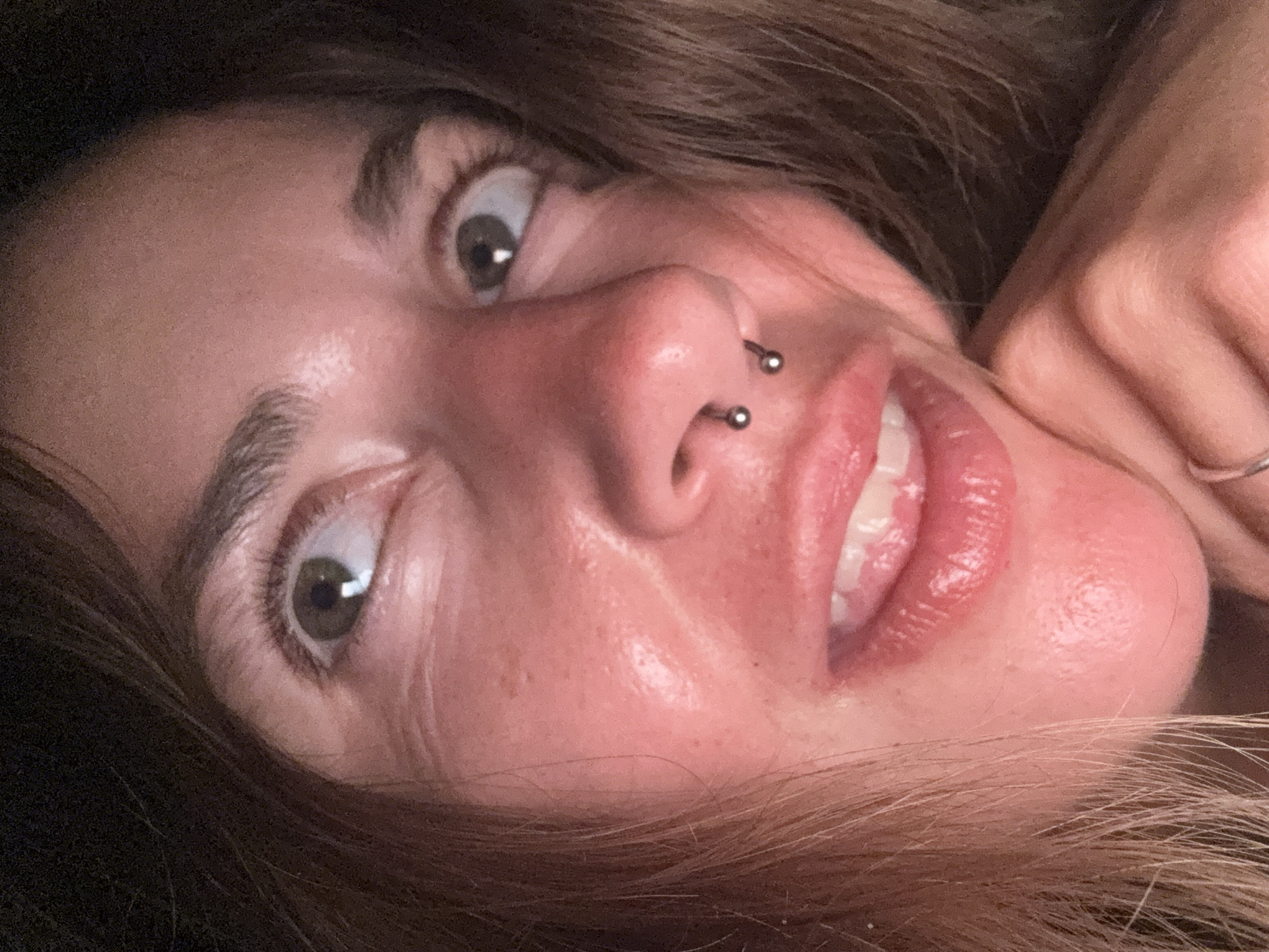 A close-up, low-angle, indoor photograph shows a young woman's face from the nose up. She has brown hair framing her face and her eyes are looking upwards and to her right. She is wearing a septum piercing. Her lips are slightly parted, revealing her teeth. The woman is resting her chin on her hand, which is visible in the lower right corner of the image, and she is wearing a ring on her index finger. The lighting in the image is dim, creating some shadows on her face. The location is unknown.