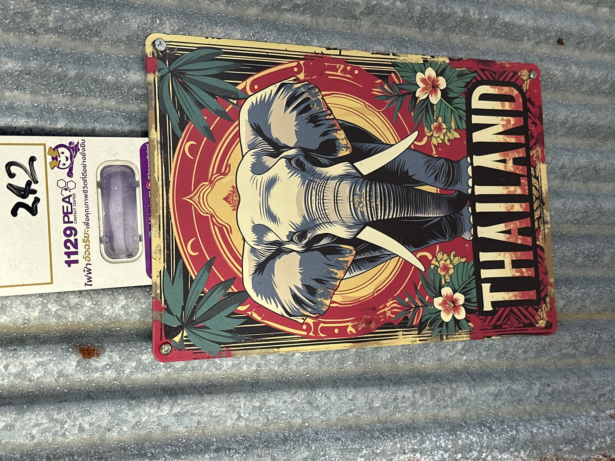 A tin sign featuring a stylized illustration of an elephant and the word "THAILAND" is attached to a corrugated metal wall. The sign has a red and gold color scheme and depicts tropical foliage. Above the elephant sign, there is a white piece of paper with Thai text and a cartoonish mascot. The metal wall has visible rust spots. The image is taken in an outdoor environment, possibly a street or market in Thailand.