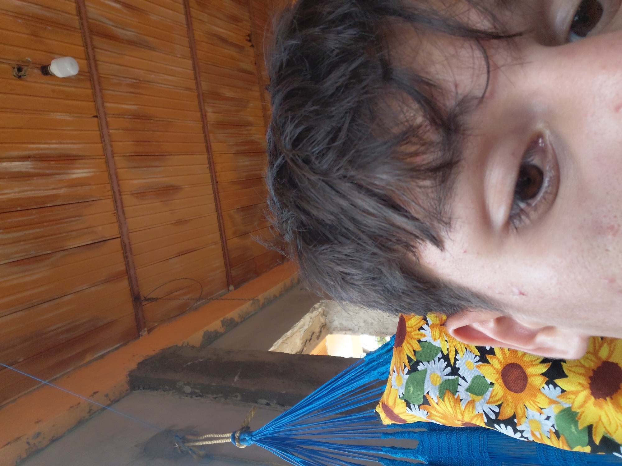 The image captures a close-up of a person's face, specifically the right eye and part of the forehead, in an outdoor setting. To the left, a blue hammock with a sunflower-patterned pillow is visible. The background features a wooden ceiling and a concrete structure, suggesting a rustic or unfinished outdoor living space. A light bulb hangs from the ceiling. The image appears to be taken in Santa Ana, Venezuela, likely a residential area given the hammock and outdoor decor. The lighting suggests it is daytime.