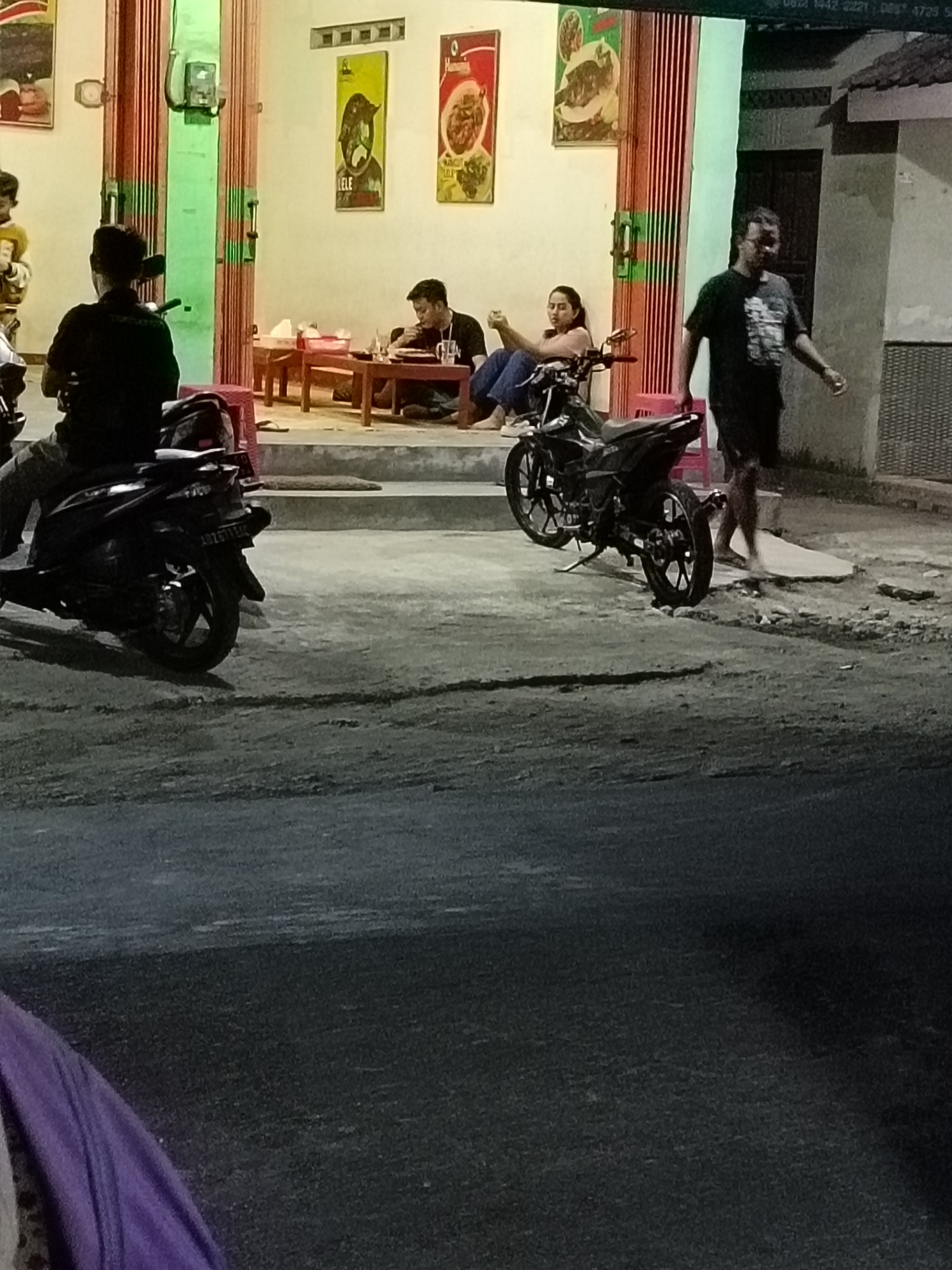 The image depicts a scene outside a food stall in Manisrenggo, Indonesia, at night. Several individuals are present. In the foreground, a person is seated on a dark-colored motorcycle. To the left, another person wearing a dark shirt is also seated on a motorcycle. Further back, two people are seated at a table. One, a man wearing a dark t-shirt, is eating. A woman in a light-colored top and dark pants is sitting with her hands clasped. To the right, a man in a black t-shirt and dark shorts is walking away from the stall. Two black motorcycles are parked near the stall. The stall itself has a light-colored exterior with vertical green and red striped panels and artwork displayed on the walls. The ground is a paved surface. The lighting suggests it is nighttime, with artificial light from the stall illuminating the area.
