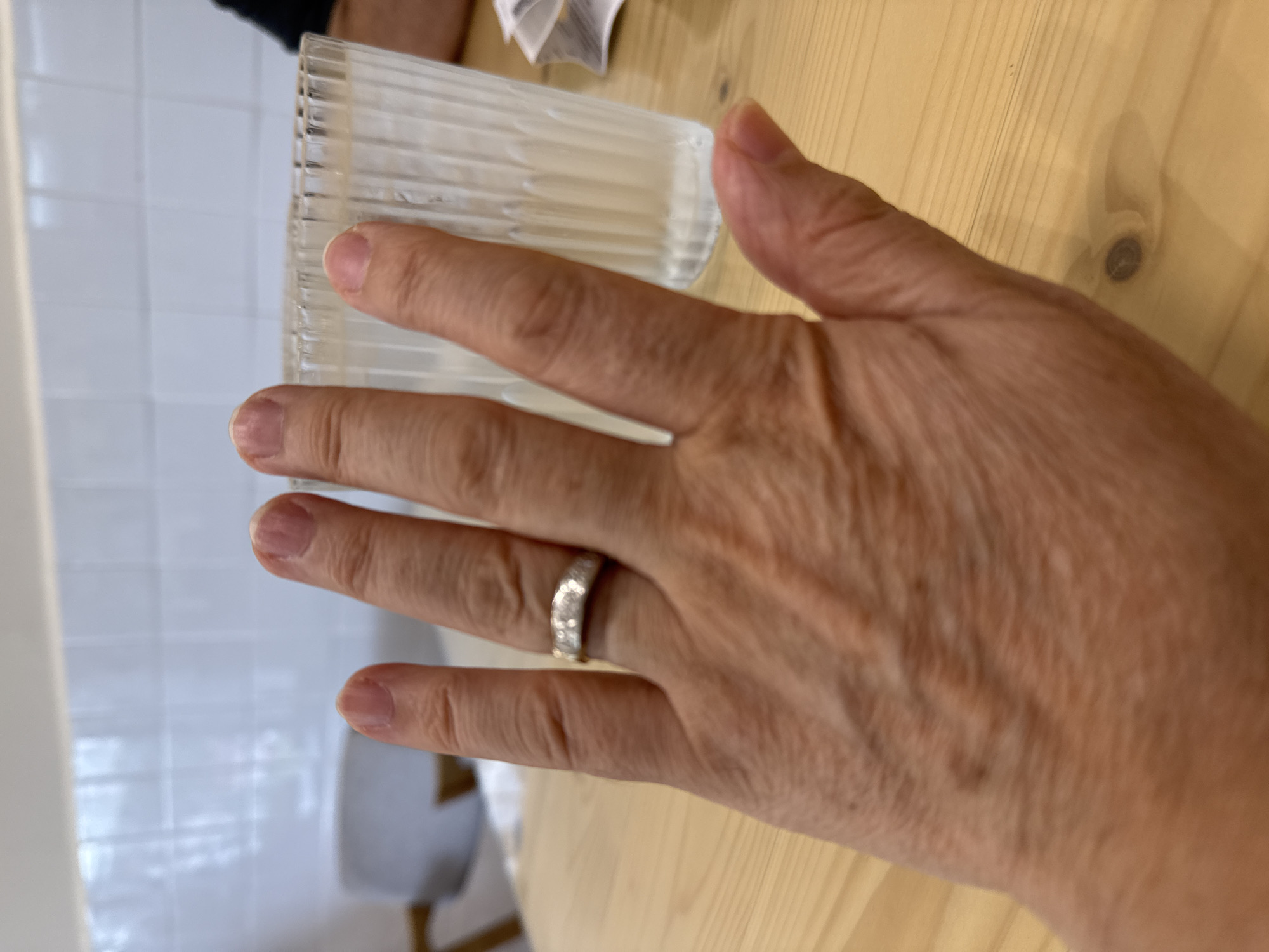 A person's hand, with a diamond band on the ring finger, is held up in front of a glass of water on a wooden table. The background shows a white tiled wall and a blurred seating area. The image appears to be taken indoors, possibly in a cafe or restaurant, but there is no specific indication of the city of Kuala Lumpur, Malaysia.