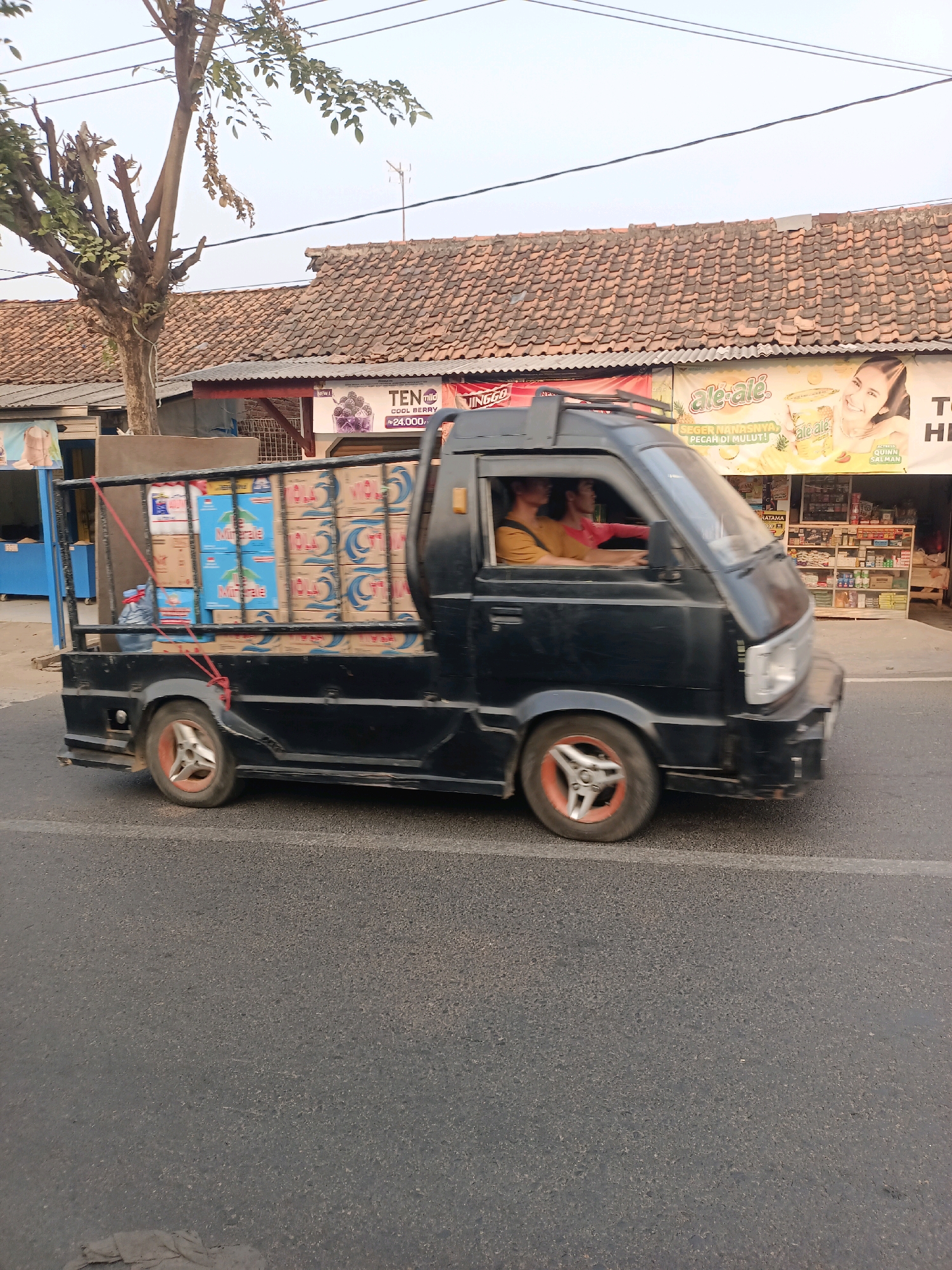A black pickup truck is driving on a paved road in Karawang, Indonesia. The truck bed is filled with cases of bottled water, secured with red ropes. Two people are visible in the cab of the truck; a driver wearing a yellow shirt and a passenger in a red shirt. In the background, there is a row of buildings with tiled roofs, one of which appears to be a small store with shelves stocked with various goods and promotional posters. A tree with sparse leaves stands to the left of the buildings. The sky is overcast.