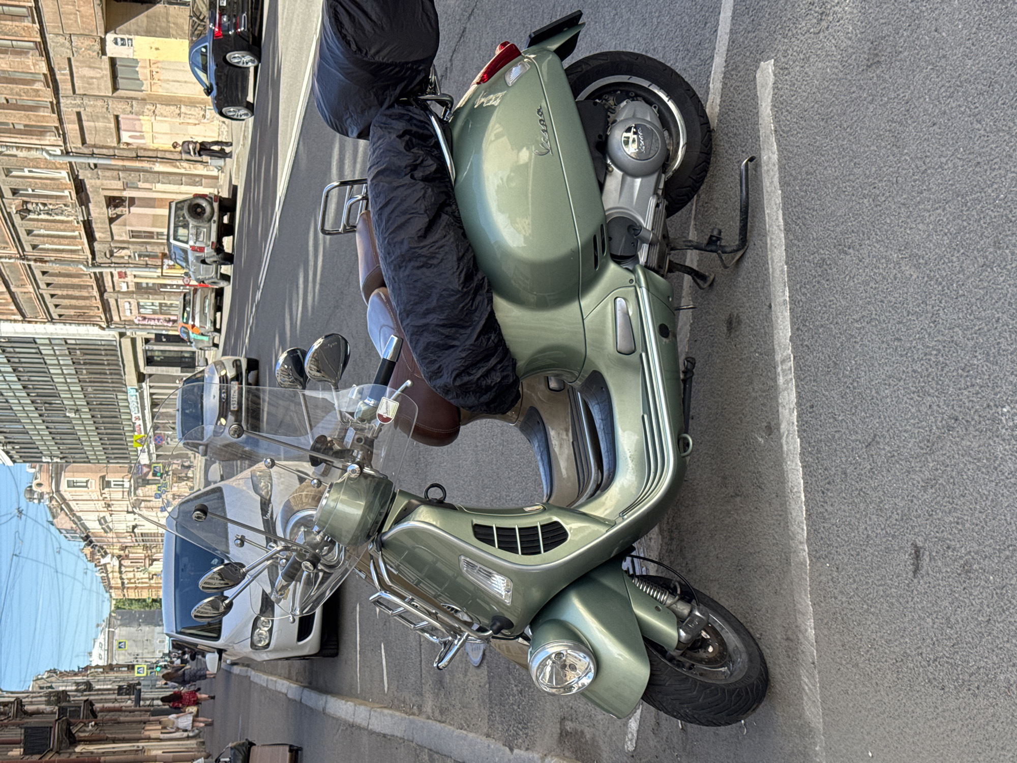 This image captures a street scene in Saint Petersburg, Russia, under a partly cloudy sky with visible patches of blue. The primary subject is a sage green Vespa scooter, likely a GTS model, parked on the right side of the street, facing away from the background. It features a brown leather-like seat, chrome front and rear luggage racks, and a windscreen. Black, wrinkled fabric covers are placed over both passenger and rider sections of the seat and possibly luggage on the rear rack. The scooter's front wheel is turned slightly to the left, and its side stand is deployed.
The street is asphalt with faded white parking lines and prominent double solid white lane dividers in the distance. On the left side, a concrete sidewalk is visible, where several pedestrians, predominantly women in casual attire, walk away from the camera. A white sedan is parked along the left curb. The buildings on the left are multi-story, exhibiting classical European architectural styles with light-colored facades and numerous windows.
On the right side of the street, other vehicles are parked or visible in the distance. A silver SUV with a rear-mounted spare tire is parked further down. Behind it, a lighter-colored sedan with what appears to be a multicolored stripe on its side is visible. A black two-door coupe is parked even further down the street on the right. Russian license plates with the "178" region code (identifying Saint Petersburg and Leningrad Oblast) are visible on the silver SUV and partially on other vehicles. A man in dark clothing stands on the right sidewalk near a building. The buildings on the right side are also multi-story, with varied architectural details, some featuring ground-floor commercial establishments. Overhead electrical wires, characteristic of trolleybus or tram lines, crisscross the sky above the street. Shadows on the ground suggest sunlight from the right side of the frame.
