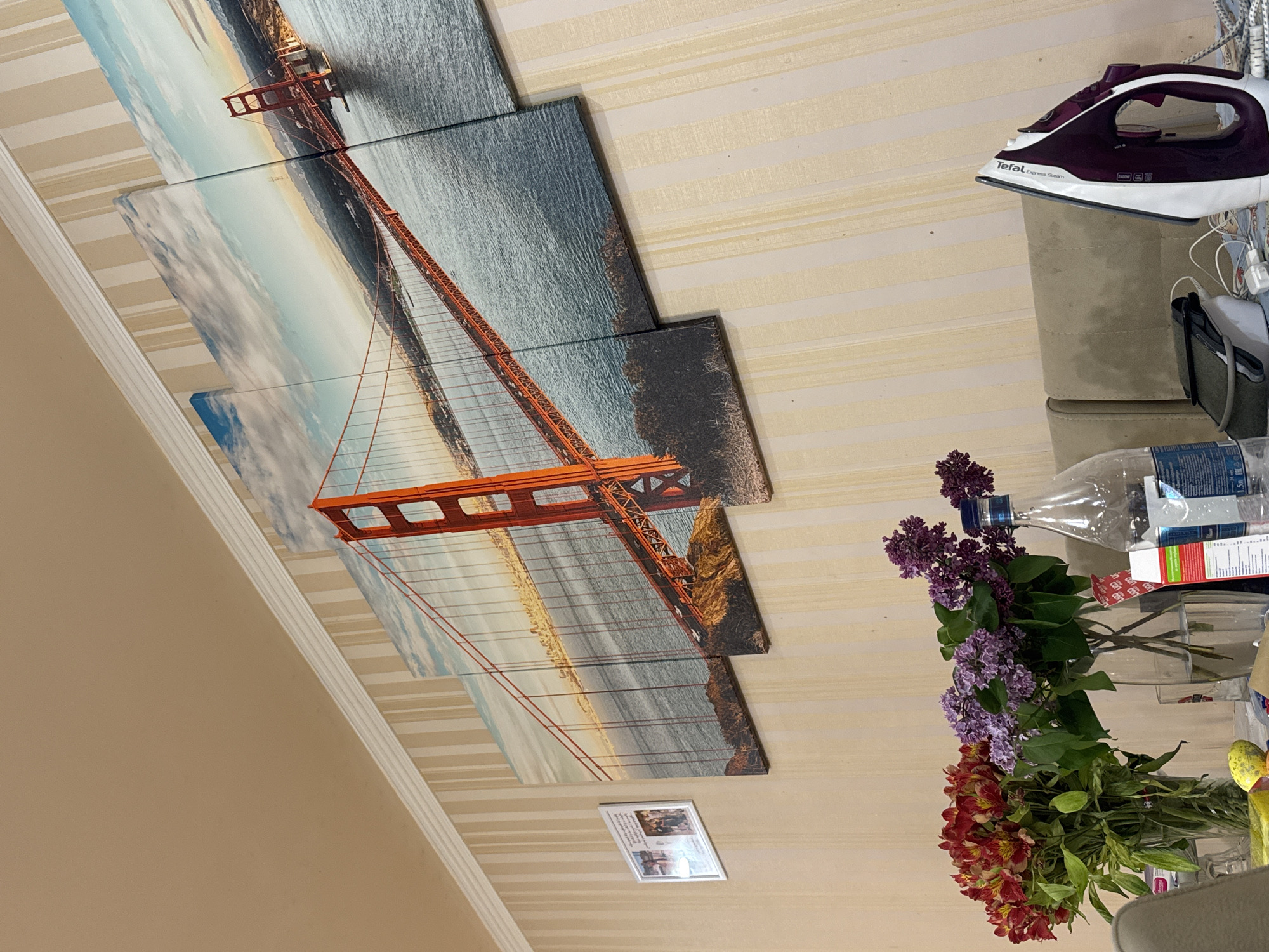 The image shows an interior space with striped wallpaper. Hanging on the wall are four panels depicting a partial view of the Golden Gate Bridge. In the foreground, a vase with flowers (lilacs and red Alstroemeria) sits on a surface. An iron sits on a piece of furniture, and near the flowers, there are transparent plastic bottles, boxes, and other items. A framed picture is present on the wall. There is no evidence of Yerevan, Armenia, or any people in the image.
