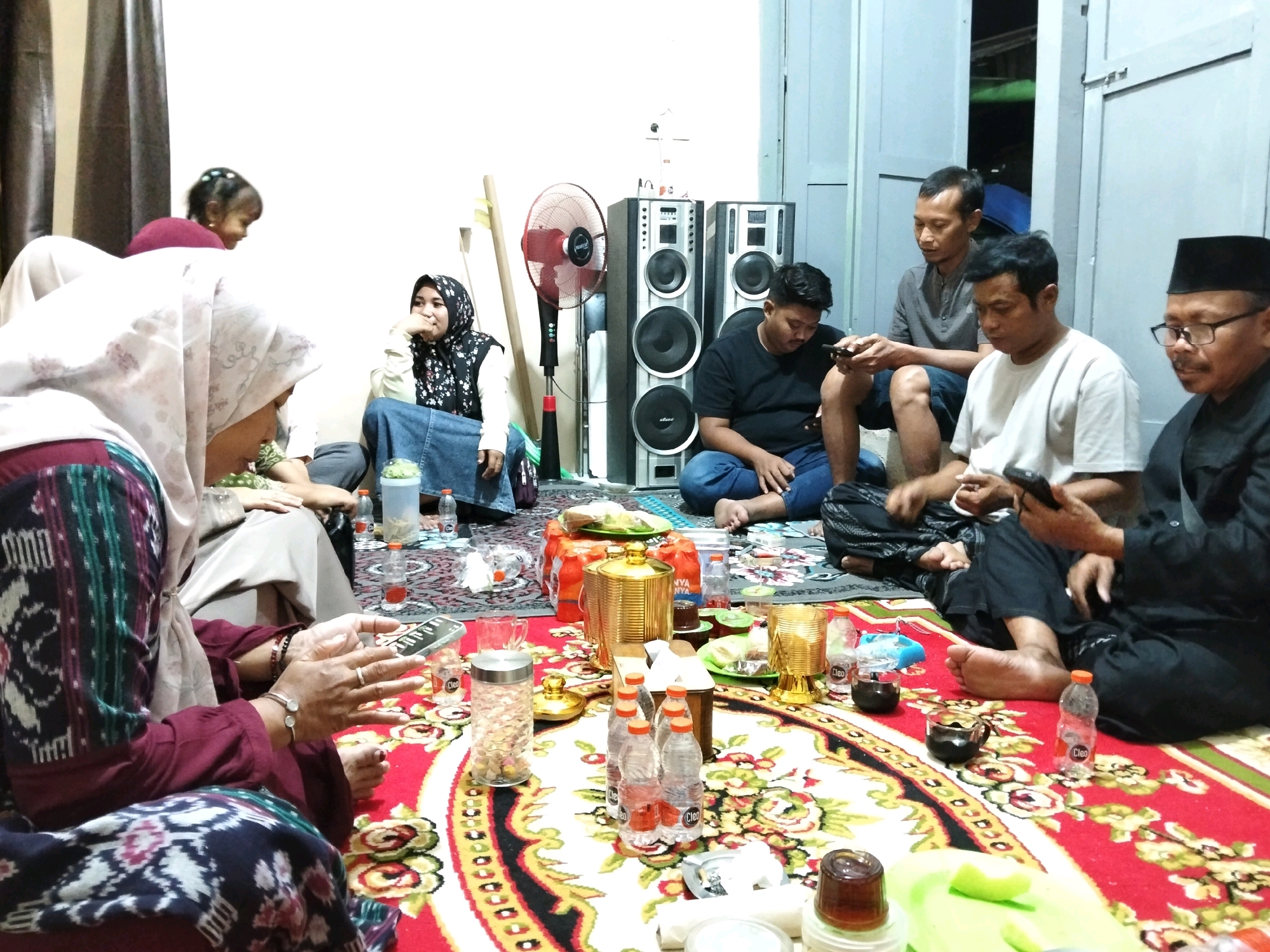 In an indoor setting, a group of people are gathered on a red carpet adorned with a floral pattern. The scene appears to be a casual social gathering, possibly in a residential area near Puskesmas Keboan, Indonesia, given the presence of traditional attire and home furnishings. Several individuals, including men and women wearing headscarves, are seated on the floor, some engaged with their mobile phones.
In the foreground, a woman in a maroon outfit and headscarf is looking at her phone. Next to her, a large gold-colored container sits on the carpet, surrounded by bottles of water and cups. In the background, a red standing fan is visible, along with two large speaker cabinets. The overall atmosphere suggests a relaxed and informal occasion, with food and drinks laid out on the carpet for consumption. The lighting is dim, indicating an evening or indoor gathering. The presence of speakers and a fan suggests a focus on entertainment and comfort within a domestic space.
