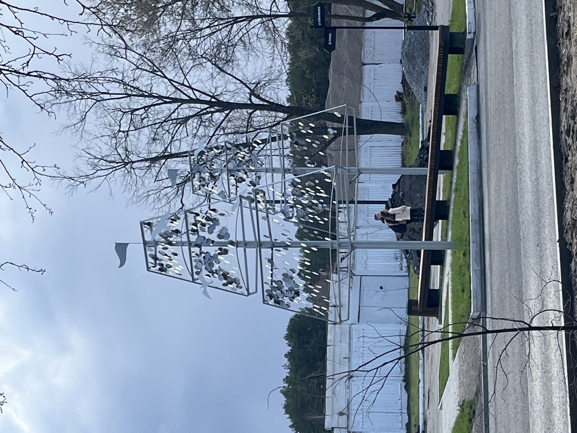 An outdoor scene in Chaikovsky, Russia, features a unique, modern art installation in the foreground under an overcast sky. The large, sculptural piece is a metallic framework, possibly representing a stylized ship or a geometric tree, adorned with numerous small, reflective, leaf-like or bird-shaped elements that catch the light. A small white flag tops the structure.
To the right of the artwork, a person is seated on a dark wooden bench with metal supports. They are dressed in light-colored outerwear over dark clothing and appear to be looking downwards or into the distance, contributing a sense of quiet contemplation to the scene. The bench is situated next to a paved pathway or road that runs along the right side of the frame.
The environment suggests late autumn, winter, or early spring, as several large trees with bare branches are visible against the muted sky, particularly on the left and upper portions of the image. In the background, beyond the trees and the art installation, are white, corrugated metal structures, possibly indicating an industrial area or construction site. A patch of green grass is visible between the pathway and the background structures.
A signpost near the top right of the image provides directional cues with Russian text. One sign clearly reads "ул. Калинина" (ul. Kalinina), and another below it indicates "ул. Мира" (ul. Mira), identifying street names in the area. The cloudy, pale sky suggests daytime with diffused light.