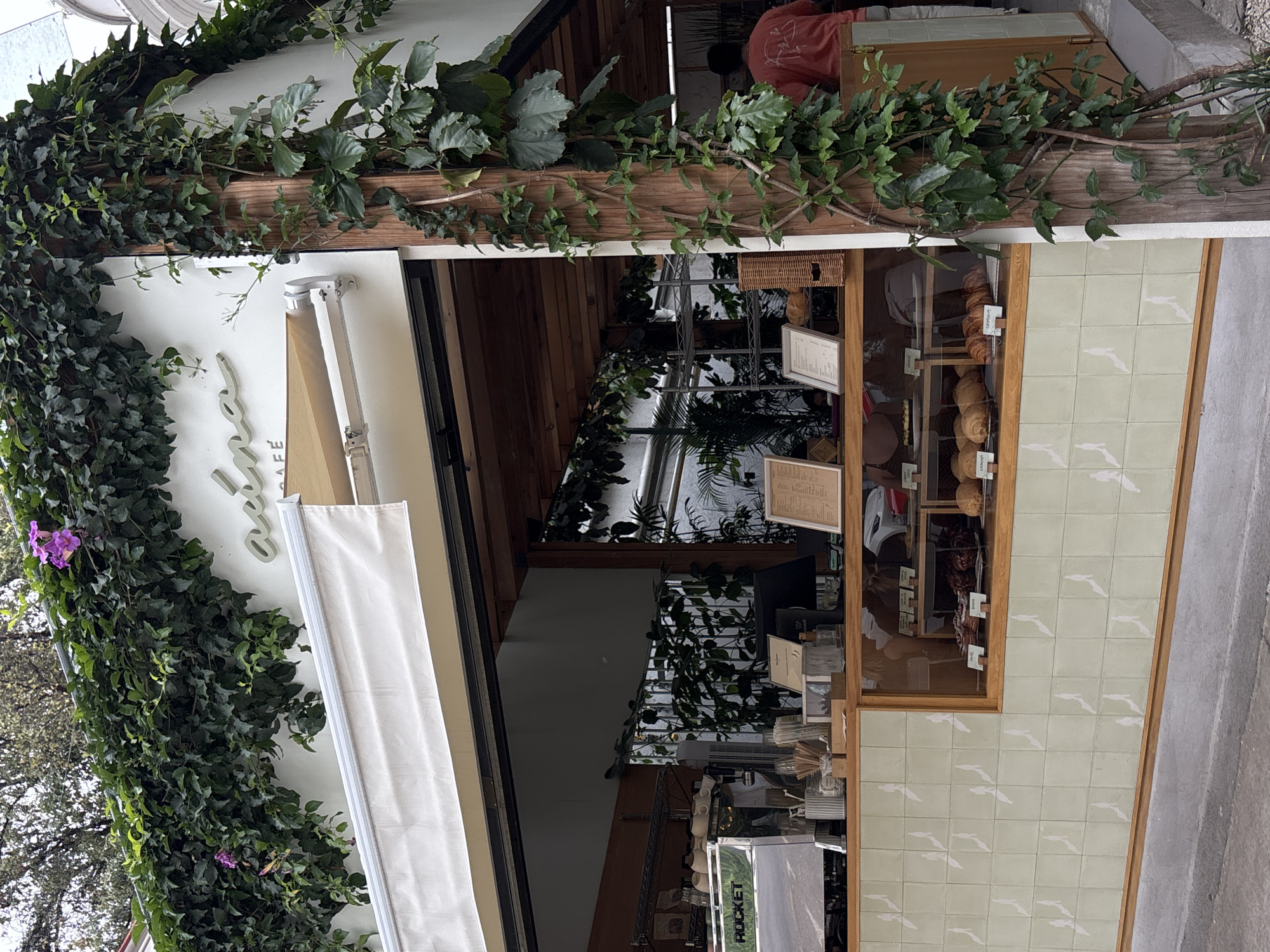 This vertically oriented image captures the inviting entrance of a cafe or bakery, likely situated on a street in Mexico City. The storefront is heavily adorned with vibrant green foliage, including various leafy vines, that cascades over a wooden beam running along the top and down the left side of the facade. Clusters of small purple flowers punctuate the dense greenery.
A partially retracted white awning covers the upper part of the entrance, beneath which a stylized, light-colored sign is visible, obscured in places by the vines; parts of it suggest "Cafe." The shop features a prominent display window filled with an assortment of baked goods, including golden croissants and different types of bread, artfully arranged on wooden shelves. Small, white rectangular labels are placed in front of some items, likely indicating their names. A woven basket sits on a shelf inside the display.
The lower right portion of the facade is covered with distinctive light green square tiles, each featuring a subtle white, abstract, or bird-like pattern. Inside the shop, a person wearing a red top is visible from the back, engaged in an activity behind a counter. The interior also reveals shelves, more potted plants, and various equipment, including a machine bearing the brand name "ROCKET." Numerous small, white rectangular signs are hung throughout the interior, presumably displaying menus or other information.
The scene is illuminated by natural light, suggesting it is daytime. The overall aesthetic, with its abundant greenery and displayed baked goods, creates a welcoming and charming atmosphere for a business that appears to be open and operational.