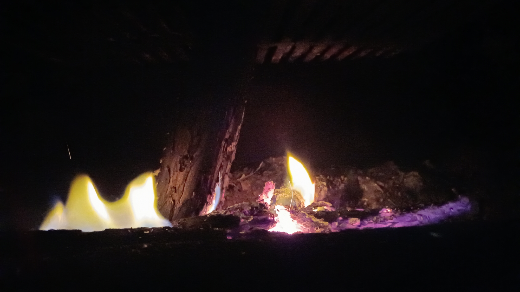 In the city of Novogusel'skii, Russia, a close-up shot captures the intense heat and light of a fire. The flames, primarily yellow and white, with hints of blue and purple at their base, dance and flicker against a dark, almost black background. The fire illuminates a rough, textured surface that appears to be burning wood or embers, with pieces of bark and charred material visible. Above the flames, a dark, slatted structure, possibly a grate or the underside of a grill, is silhouetted against the glow. The overall impression is one of raw, elemental energy, with the darkness emphasizing the brightness and warmth of the fire. There are no people present in the image.