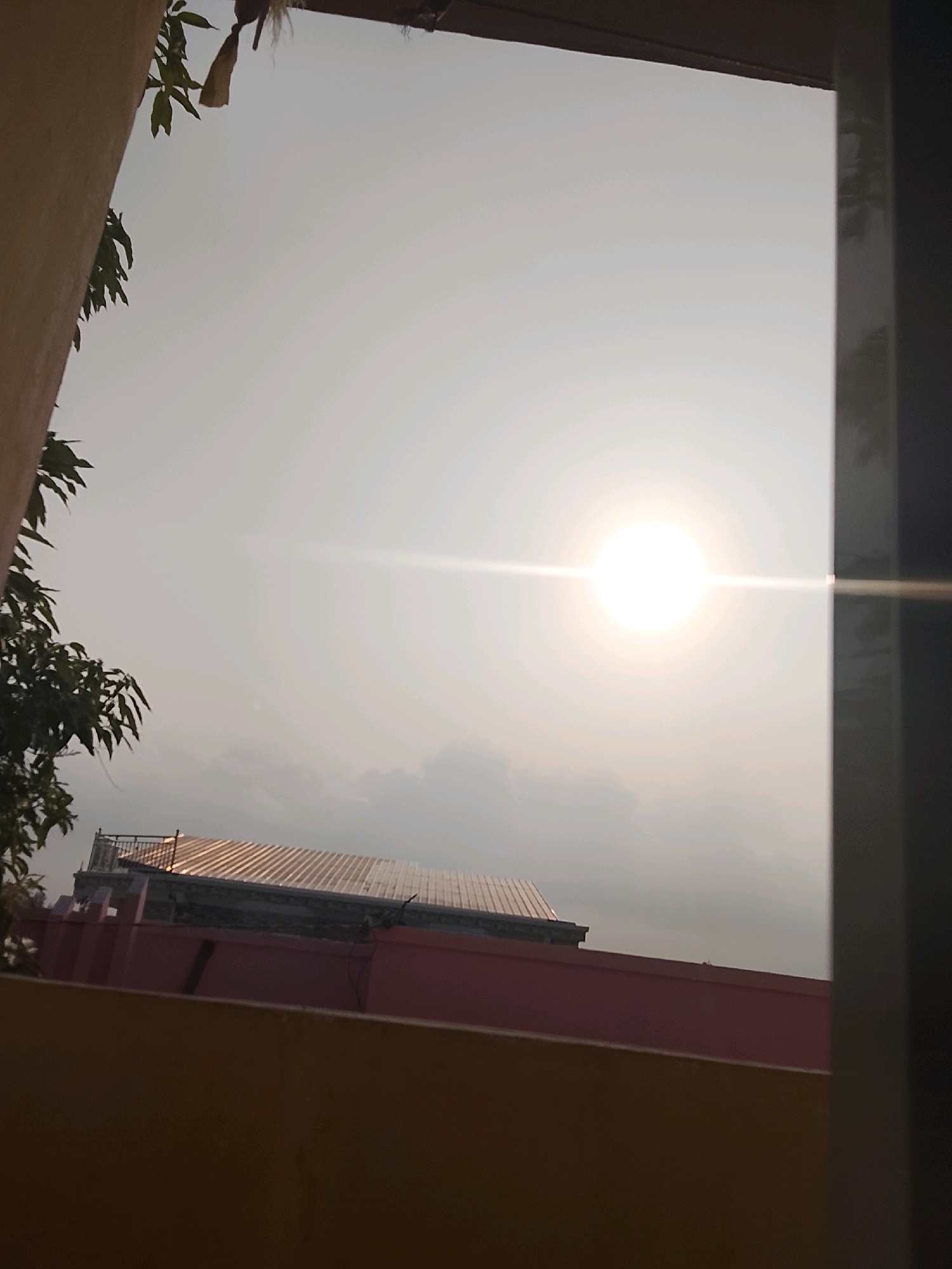 The image shows a bright sun in a hazy sky, with a light flare emanating horizontally from its center. In the lower portion of the frame, parts of a building are visible, including a red wall and a roof with a corrugated metal surface. To the left, a tree with green leaves is partially obscuring the sky. The scene appears to be an outdoor view, possibly from a window or balcony, overlooking rooftops in a tropical or sub-tropical urban setting. The quality of light suggests it is daytime, potentially late afternoon or early morning, given the haze. The setting is consistent with a location in Tanjung Balai Karimun, Indonesia, characterized by its coastal urban environment and tropical climate. There are no people or discernible actions depicted in the image.