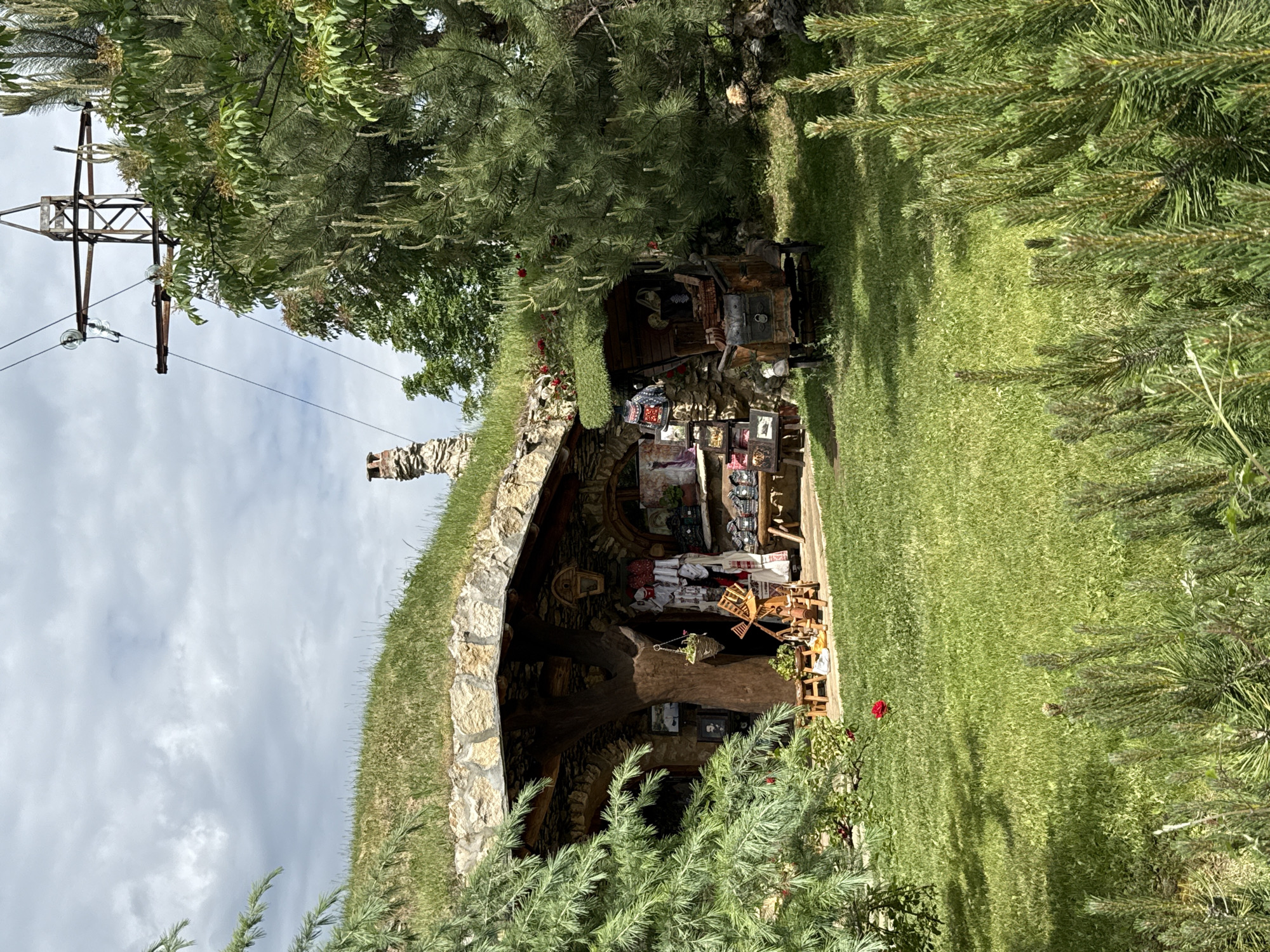 The image depicts the Eco Path "Golden Panega" near Lukovit, Bulgaria. A structure with a stone exterior and grass-covered roof is visible. The interior space is filled with various items, including what appear to be decorative pieces, artwork, and furniture. In the surrounding environment there is lush green grass and various trees. Overhead, a power line is visible against a cloudy sky.