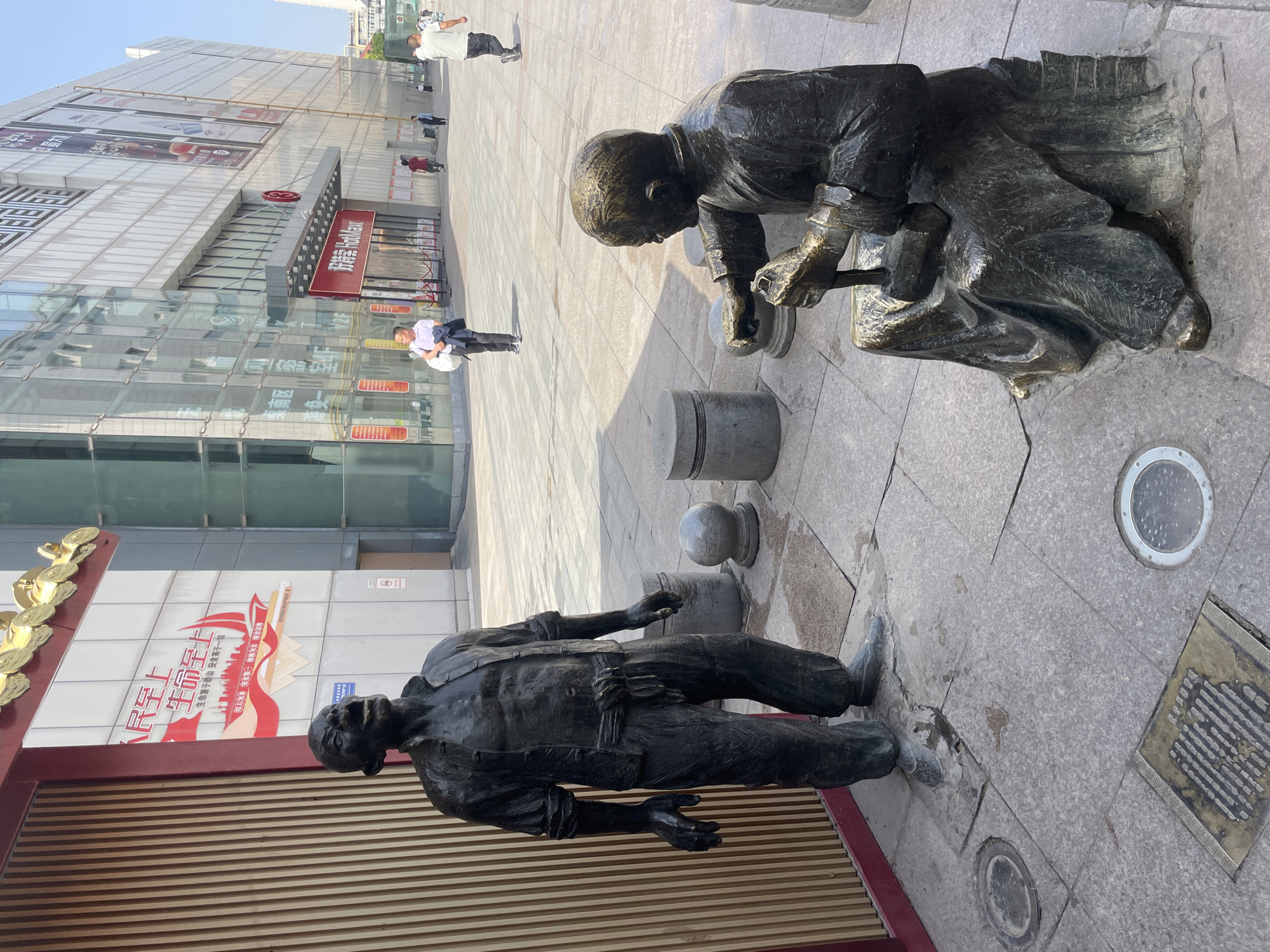 The photo shows two bronze sculptures of people interacting in Xincheng District, China. One sculpture depicts a person seated and hunched over, appearing to be working on something in their hands. The other sculpture is of a person lying prone on the ground.
Behind the sculptures, there is a building with a glass facade and signs in Chinese, including "人民至上" (People First) and "生命至上" (Life First). A red banner with white text indicates "好特卖 HotMax," likely a store name. People are walking in the background. The ground is paved with stone tiles. The photo seems to be taken at an angle, giving the scene a slight tilted perspective.