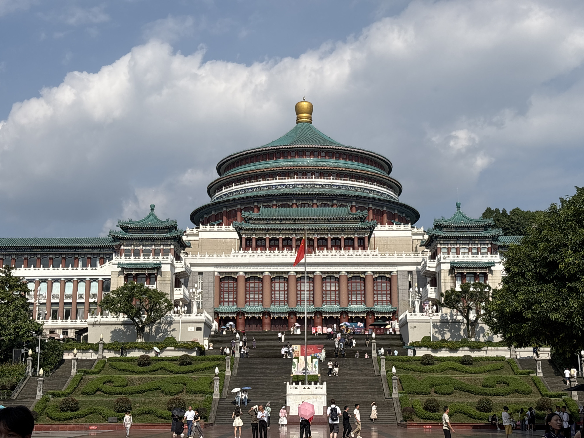 The image shows the Great Hall of the People in Yuzhong District, Chongqing, China, on a bright, cloudy day. The grand building, a prominent landmark, features traditional Chinese architecture with multiple tiered roofs covered in green tiles and a golden dome atop the central rotunda. A Chinese flag flies from a tall flagpole in front of the building.
A wide, grand staircase leads up to the main entrance, lined with people walking and standing. The stairs are flanked by meticulously manicured green hedges arranged in decorative patterns, interspersed with small trees and shrubs. Stone planters with ornate lamp posts are also visible along the staircase.
Numerous people are scattered throughout the scene, some ascending or descending the stairs, others gathered in groups at the bottom. Many are holding umbrellas, suggesting it might be sunny or there is a possibility of rain. The atmosphere appears to be one of tourism and public gathering. The background reveals more of the building's architectural details, including rows of windows and decorative columns, as well as lush green trees bordering the property. The sky is a mix of blue and large, puffy white clouds.