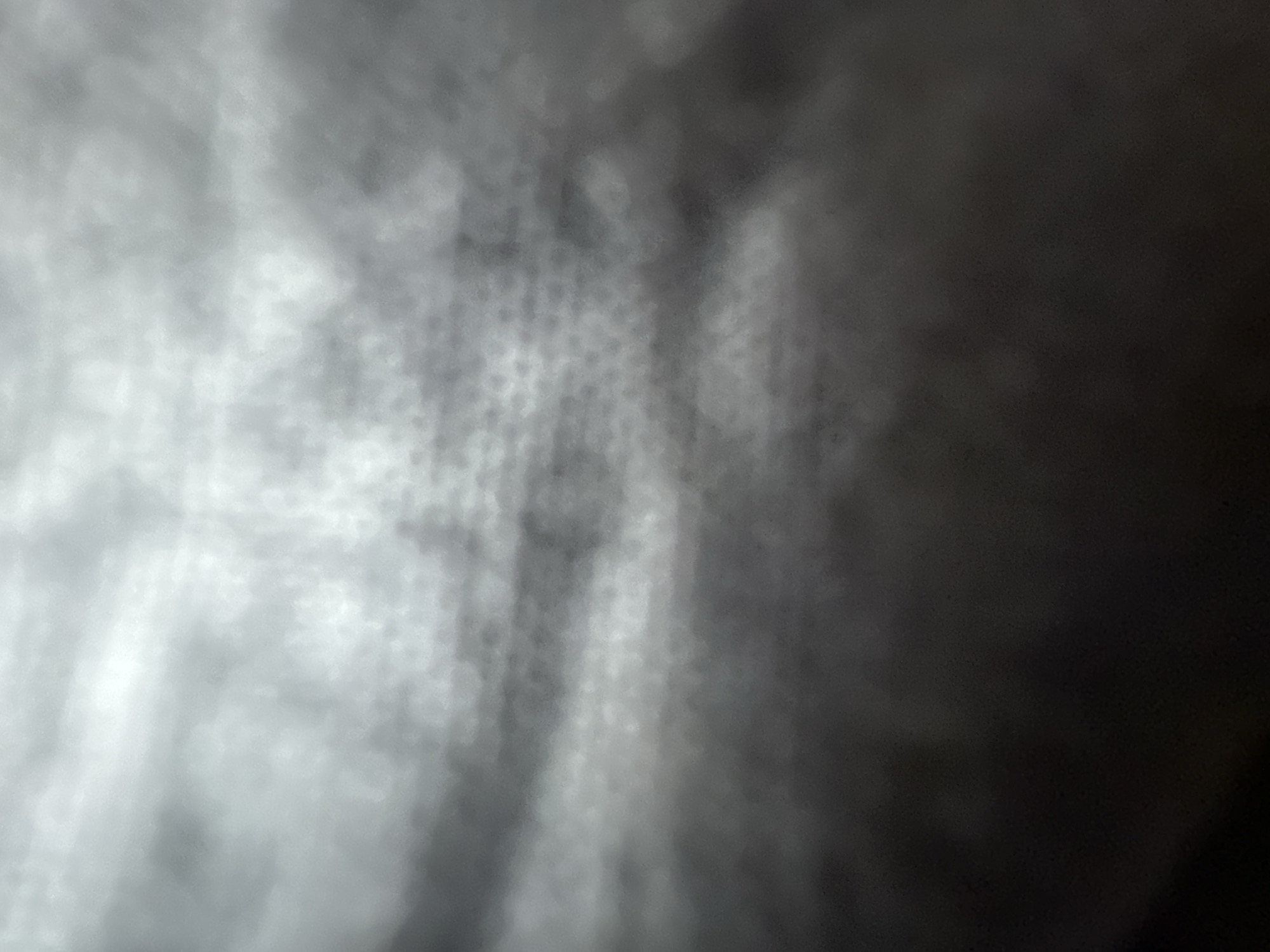 The image is an abstract, blurred photograph with a predominantly grayscale palette. It appears to be an extreme close-up of a textured surface, possibly fabric or a wall, with a pattern of small, circular indentations visible in some areas. The lighting is dim and uneven, creating a gradient from dark gray at the bottom to lighter grays and a very bright, almost white area at the top. There are no discernible people, objects, actions, or interactions. The image does not contain enough information to determine a specific location within the city of Belgrade, Serbia.