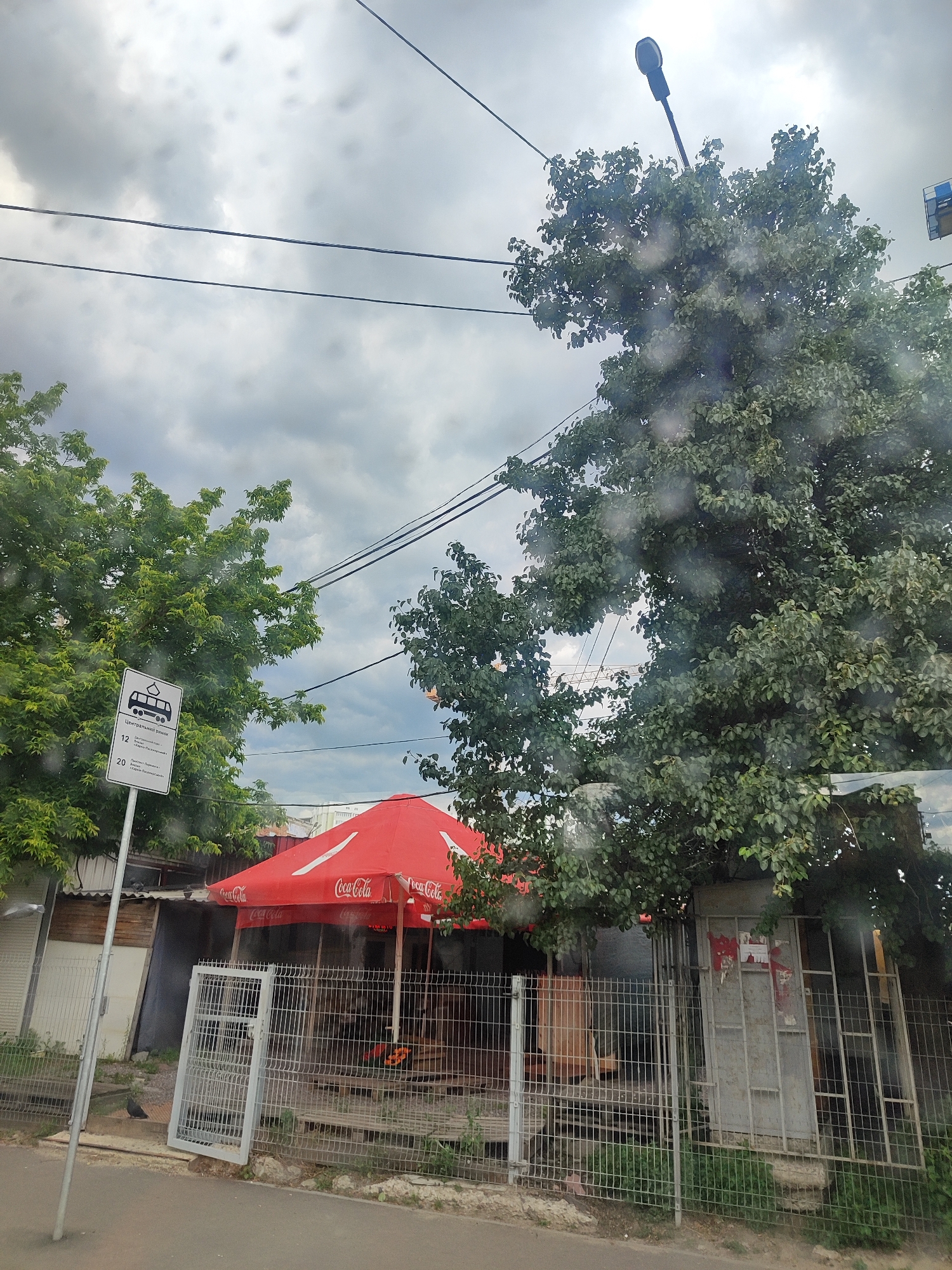 The image depicts an outdoor scene with a cloudy sky overhead. In the foreground, a paved surface transitions to a sidewalk or grass area bordered by a metal fence. Behind the fence, there is a small building or kiosk with a red umbrella, branded with "Coca-Cola." A bus stop sign with Cyrillic text and route numbers is visible to the left, near a tree with green foliage. A street lamp is also visible in the background, perched on a pole. The overall impression is of a modest urban or suburban setting, possibly near a transportation hub or commercial area.
