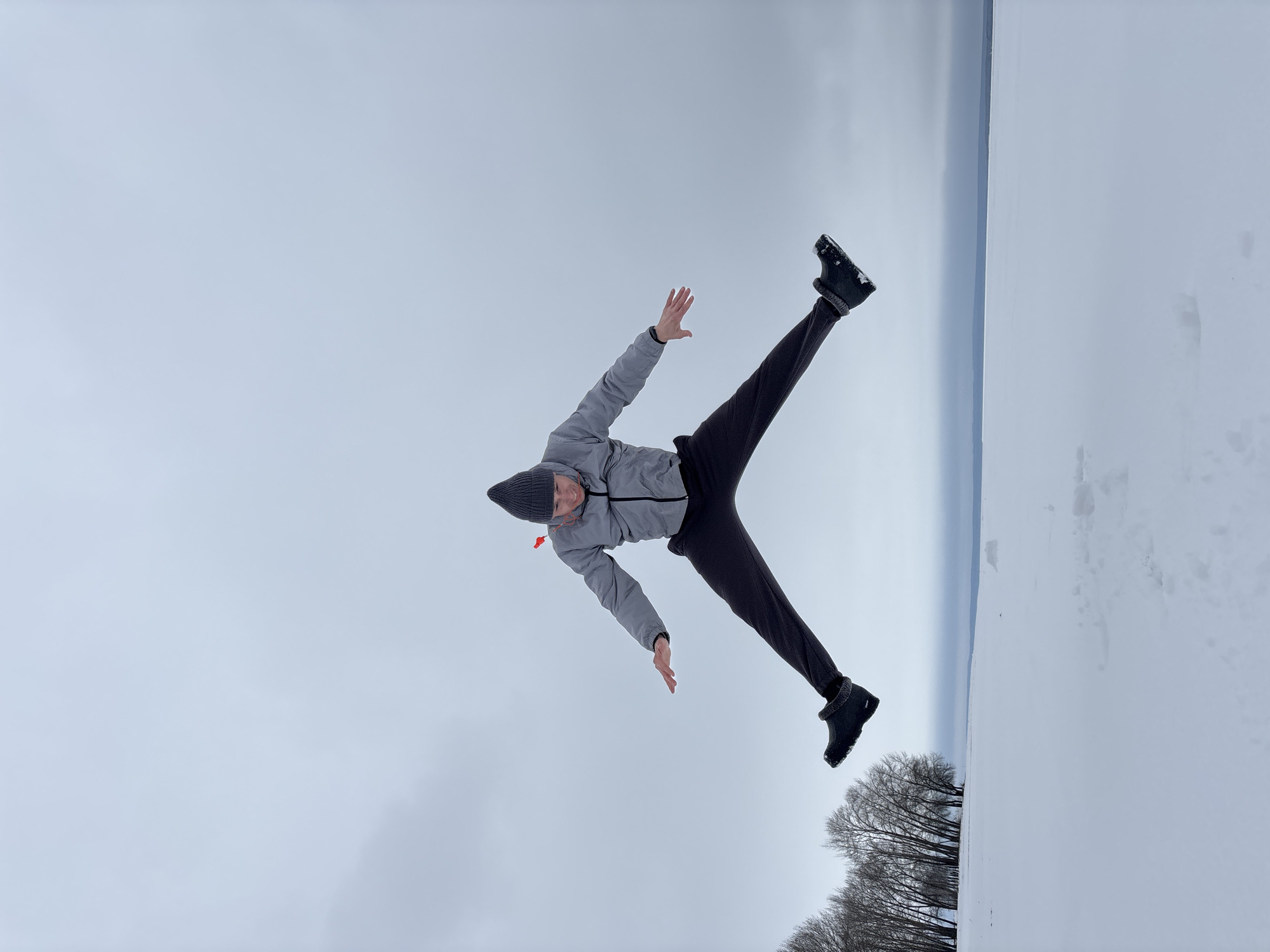 A person is lying on their back in the snow near a lake in Noviy, Russia. The individual wears a gray jacket, dark pants, a gray beanie, and dark boots. Their arms are extended to the sides, and legs are spread apart, forming a star-like shape on the snowy ground. The sky is overcast. In the background, a line of trees separates the snow-covered ground from what appears to be a frozen lake or body of water. The image is taken from a low angle, giving the impression that the person is floating against the backdrop of the sky and lake. The photograph is rotated clockwise by approximately 90 degrees.