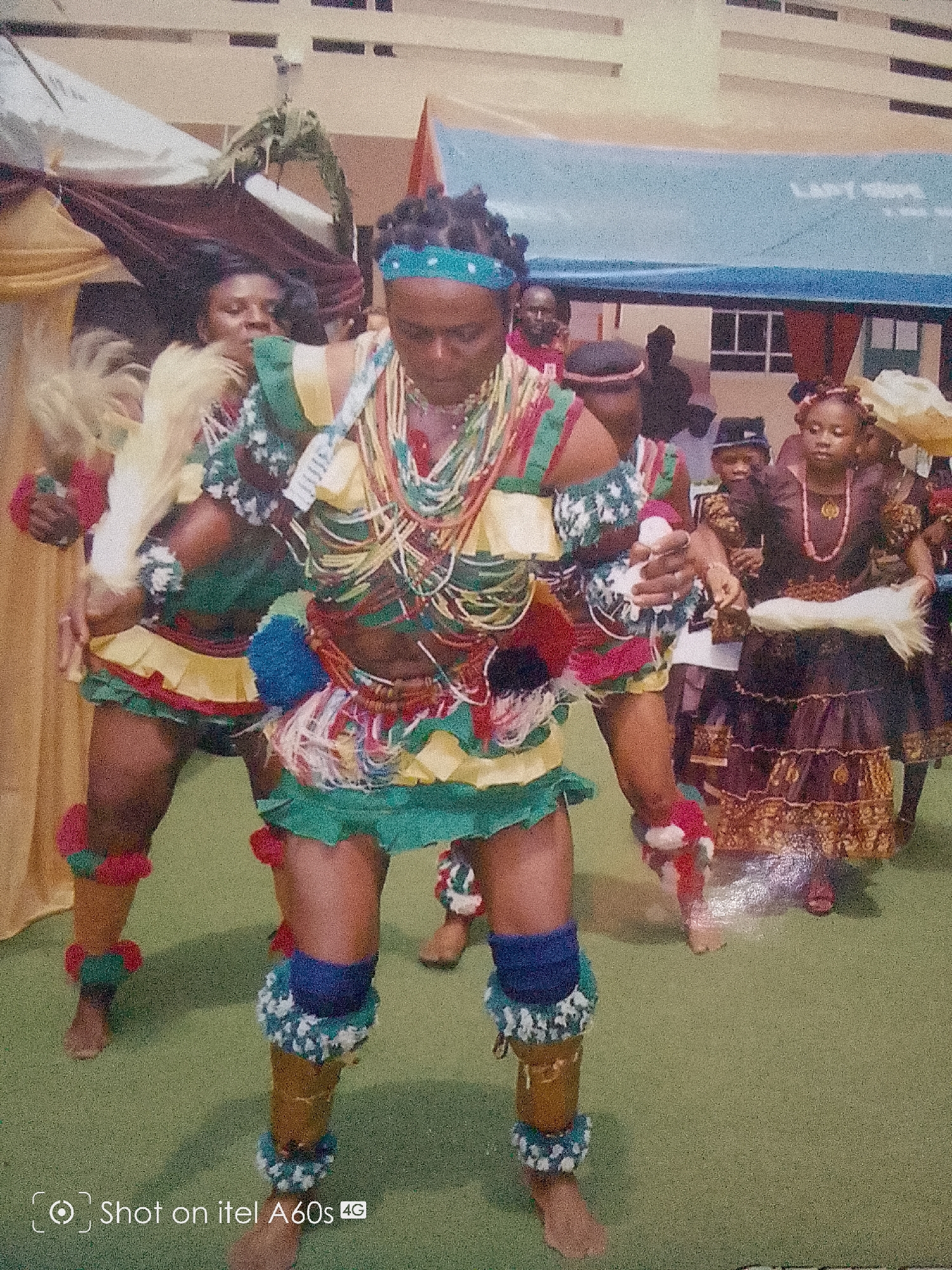 In Idimu, Nigeria, a group of people are gathered, seemingly for a cultural event. Several individuals, predominantly women, are dressed in traditional attire featuring green, yellow, and red colors. The individuals are adorned with beaded necklaces and leg decorations. The setting appears to be outdoors, possibly on a green grass lawn. A building is visible in the background. The scene suggests a celebration or performance within a cultural context.