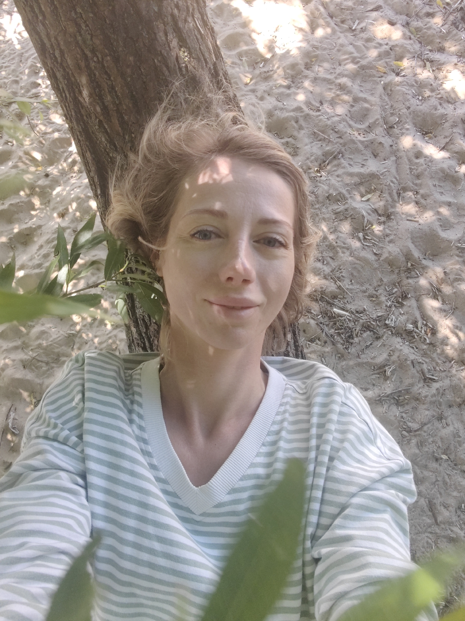 A young adult female with blonde hair, fair skin, and blue eyes is taking a selfie while leaning against a tree. She is wearing a light green and white striped V-neck sweater. The background consists of sand with scattered dry twigs and leaves, illuminated by dappled sunlight filtering through foliage, suggesting an outdoor, possibly park or beach-like setting. There is no indication of the location being in Saint Petersburg, Russia.