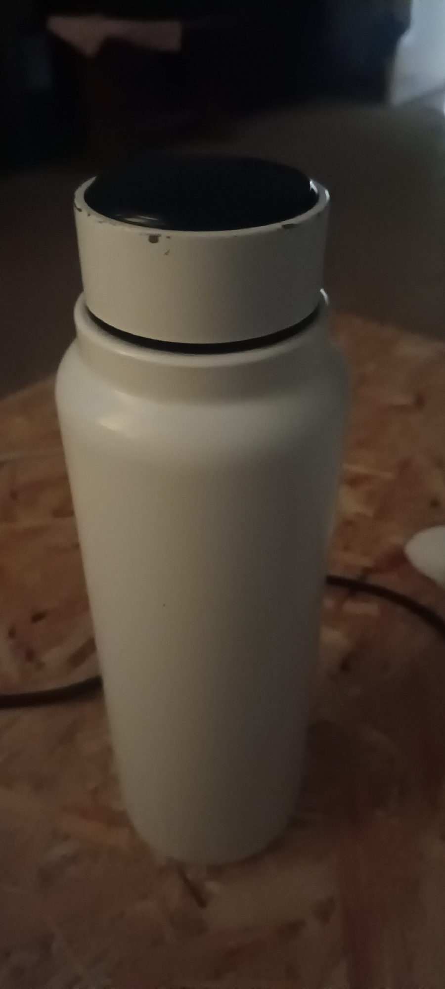 A white, cylindrical thermos with a black lid is standing on a wooden surface. The thermos is in the center of the frame. The lid has some minor scuffs and a black button in the center. A dark cable is visible behind the thermos, connecting to something off-camera. The background is dark and out of focus, showing hints of furniture. The image appears to be taken indoors. The location in Pruzhany, Belarus is not discernible from the image.