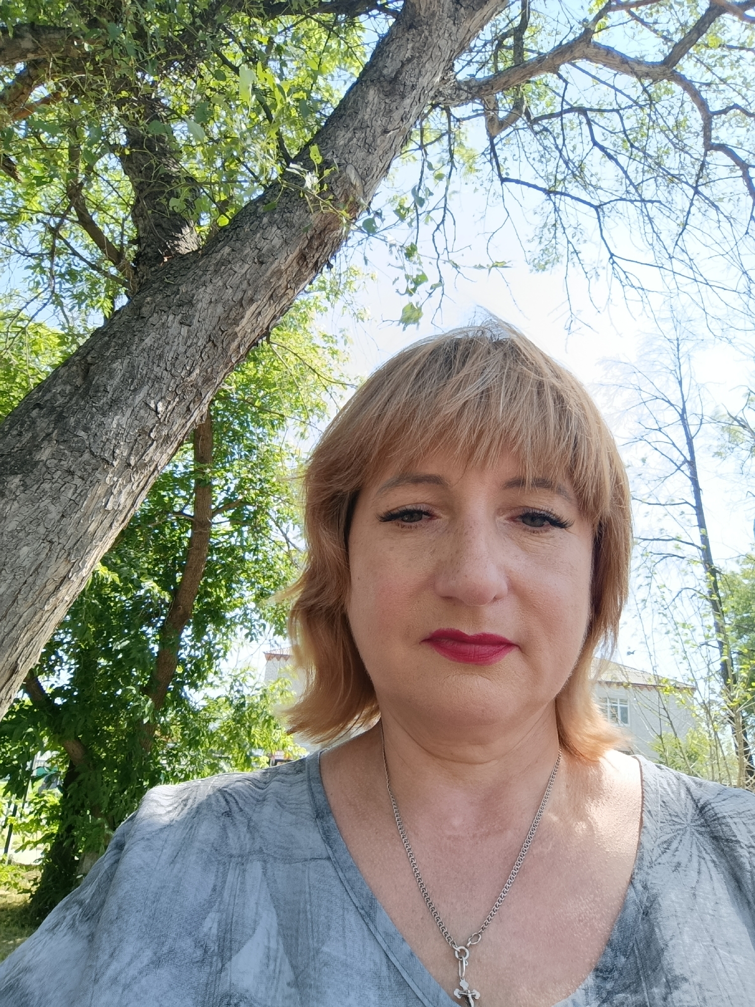 In this front-facing, medium shot taken outdoors, a woman with light blonde hair and bangs, wearing red lipstick and a grey patterned top, is positioned centrally. Her gaze is directed forward, with her facial features, including light freckles, visible. She wears a silver necklace with a pendant. Behind her, a large tree trunk dominates the left side of the frame, with its textured bark and branches extending upwards. Lush green foliage from the tree fills the background on the left, while the right side of the image shows more branches against a bright blue sky, with a glimpse of a building in the distance. The setting appears to be a park or residential area within the city of Talitsa, Russia, given the natural and built environment depicted.