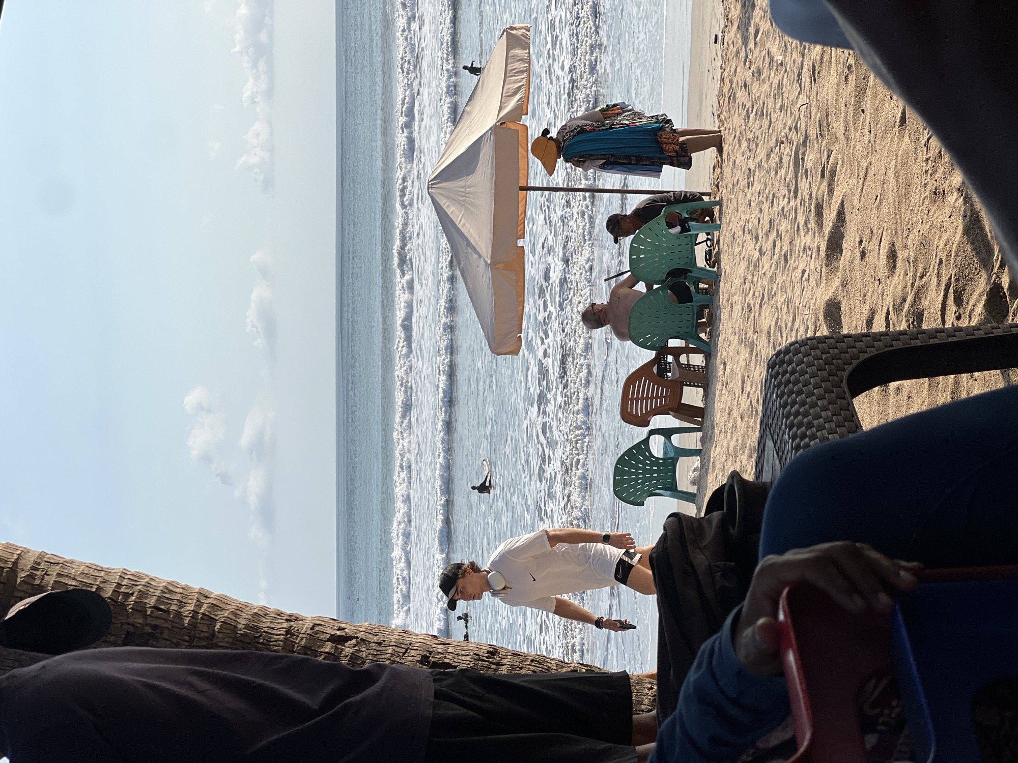 In Kuta, Indonesia, on a sunny beach, a young man wearing a white t-shirt, black shorts, and a baseball cap with headphones around his neck walks by the shore. In the background, several surfers are visible in the ocean, with waves rolling in. Closer to the shore, an umbrella shades a table with chairs where two people are seated, and another person stands beside them. The foreground shows the legs of individuals sitting on chairs, indicating a relaxed beachside setting.