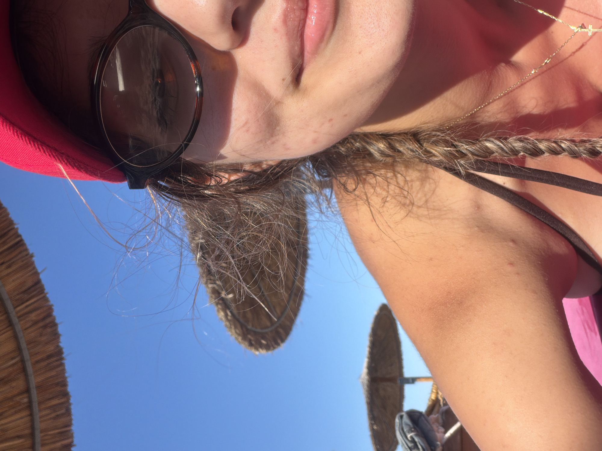 A close-up, selfie-style photograph taken outdoors during the daytime. The image features a young woman's face and upper torso, with a bright blue sky and thatched umbrellas in the background. She is wearing a red baseball cap and sunglasses, and her hair is braided. The woman appears to be at a beach or outdoor recreational area. The scene suggests a sunny day, possibly at a tourist destination. No specific landmarks are visible to identify the location within Burgas, Bulgaria.
