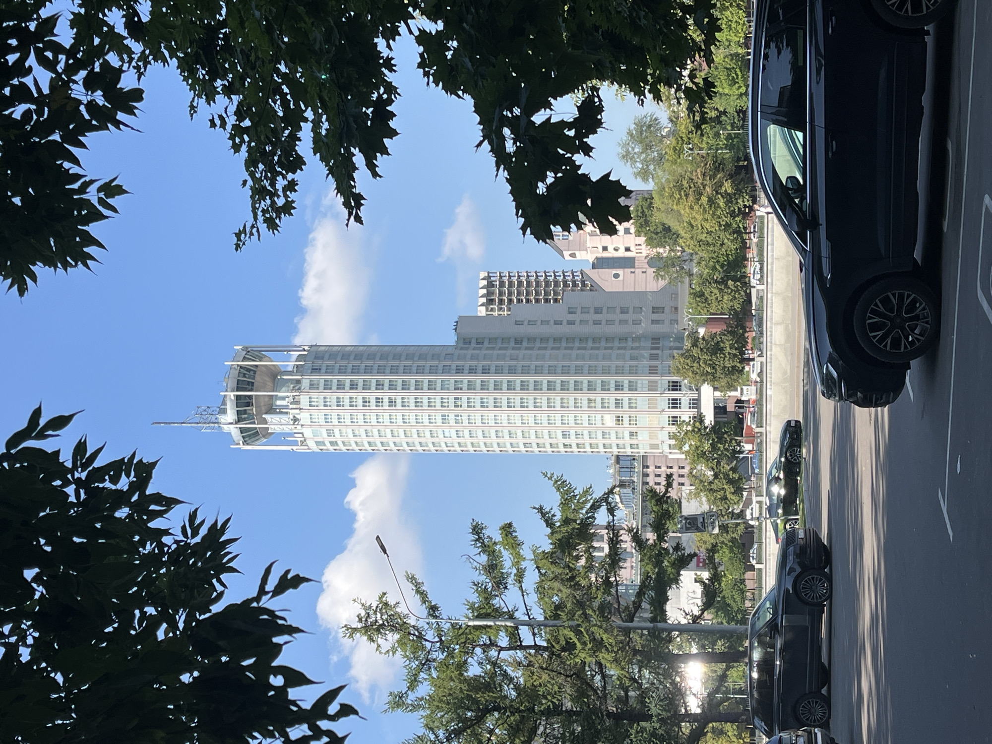 The image shows a view of the Radisson Collection Hotel, Moscow, formerly known as the Hotel Ukraina, located at Kutuzovsky Prospekt, 2/1, Building 1, Moscow, Russia. The iconic Stalinist skyscraper, a prominent landmark in the city, is visible in the background under a clear blue sky with scattered white clouds. The hotel is distinguished by its distinctive crown-like structure at the top, complete with a helipad. In the foreground, trees with dark green foliage frame the scene, partially obscuring the view. Several dark-colored vehicles, including SUVs, are parked and driving on a road in front of the hotel. A lamppost is visible on the left side of the image, and other buildings and trees are situated along the embankment of the Moskva River, which is implied to be just beyond the road. The overall scene depicts an urban environment on a sunny day.