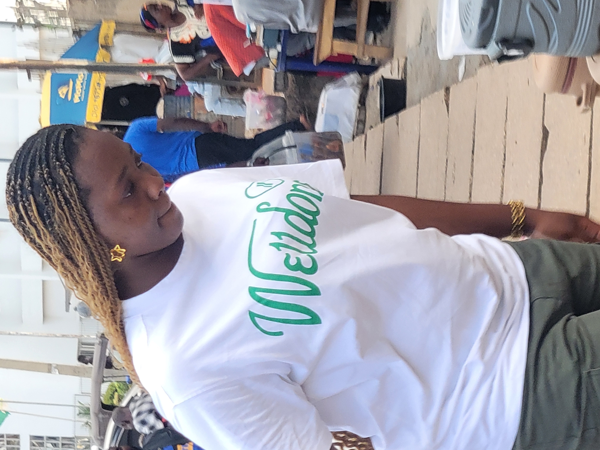 A woman with braided hair sits outdoors, wearing a white T-shirt with green lettering. She has a gold star earring and a gold bracelet. She appears to be in an urban setting, based on the building details in the background. Jos, Nigeria can be identified by the "Nomaco" sign written on a blue background. Other people are visible behind her, including one in a blue shirt. Clothing hangs from a structure in the background.
