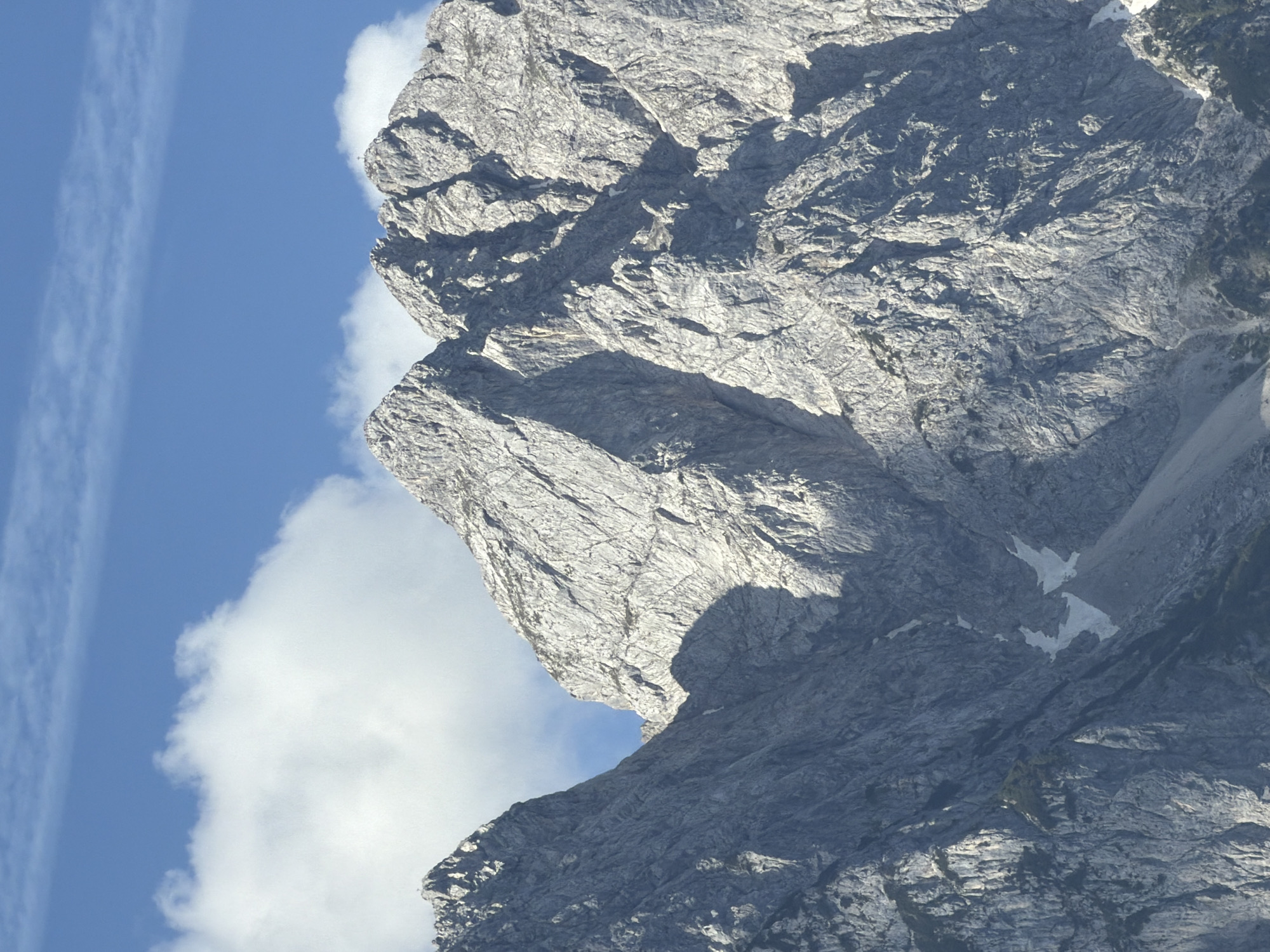 A medium shot captures a majestic mountain range under a clear blue sky dotted with wispy clouds. The mountains, composed of rugged, textured gray rock, are sharply defined, with their peaks reaching towards the heavens. Sunlight casts dramatic shadows, highlighting the craggy features and steep inclines. Patches of snow are visible in shaded crevices, contrasting with the grey rock. The lower slopes of the mountains are partially obscured by shadow and some vegetation is visible at the very bottom of the frame. The scene is devoid of any human presence or man-made structures.