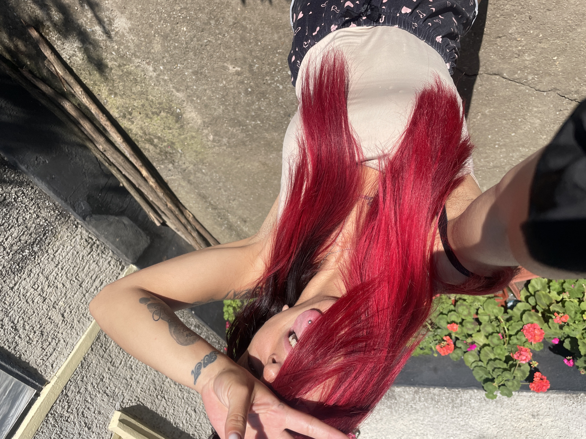 The image depicts a close-up, high-angle selfie of a person with bright red hair and a tongue piercing. The person is wearing a light-colored tank top and dark patterned shorts. Their right hand is raised, covering their eyes, and their tongue, adorned with a metal piercing, is visible. Tattoos are visible on their arm. The background shows a textured wall, a concrete ground with some cracks, and a planter with pink flowers. The scene appears to be outdoors, possibly a courtyard or patio. No specific location within Zheglartsi, Bulgaria, can be identified from the visual information provided.